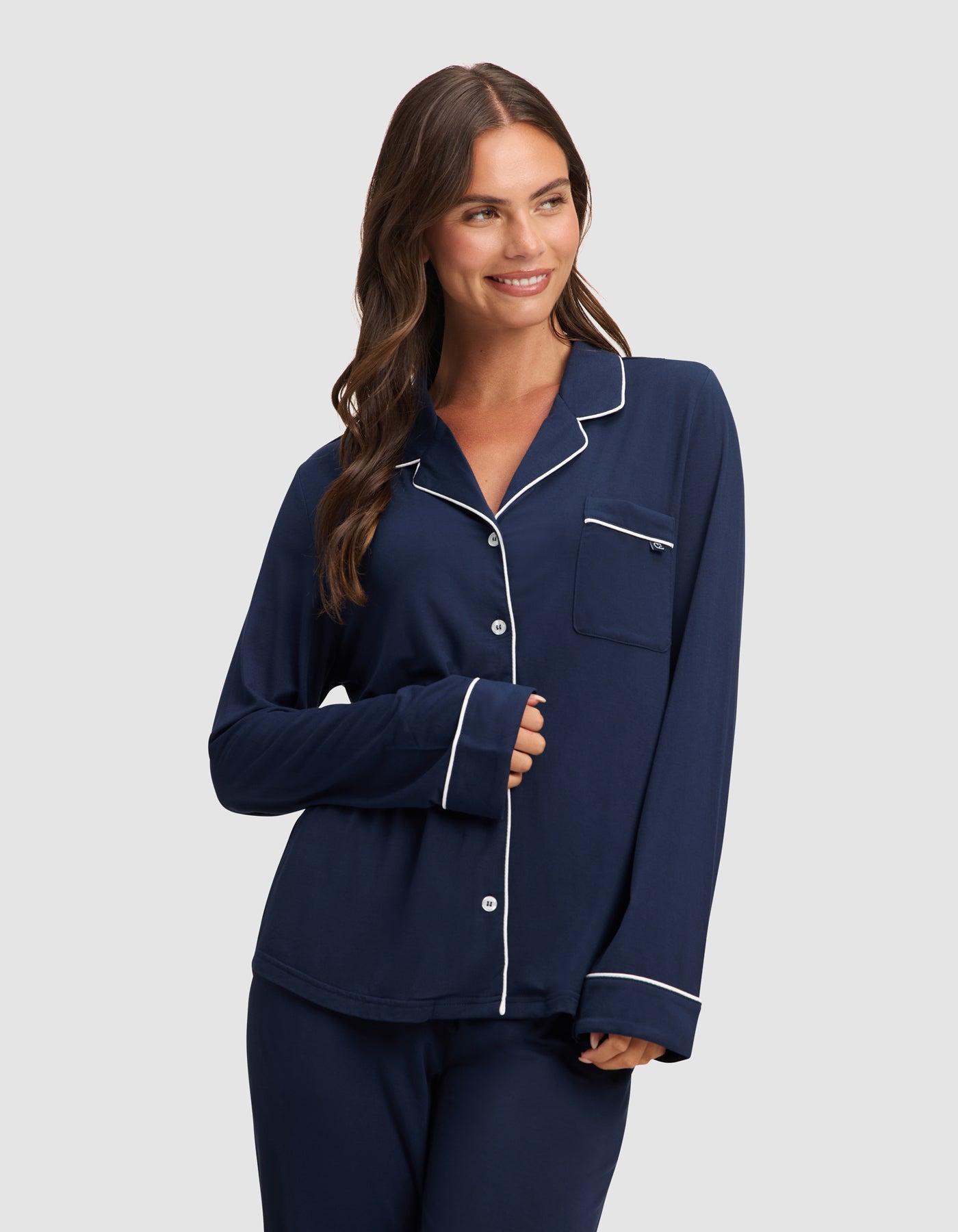 A woman with long brown hair smiles in a HIDE Women's Bamboo Stretch-Knit Long Sleeve Pajama Top, navy blue with white piping, paired with matching pants, against a plain light gray background.  
