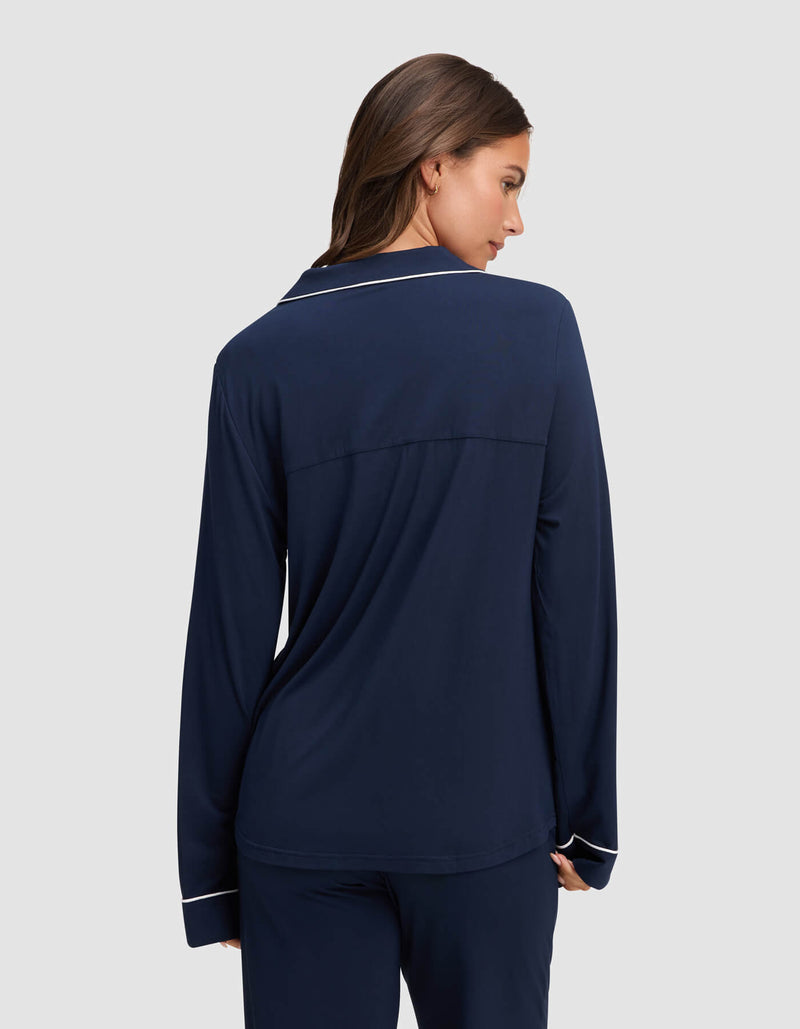 A woman with long brown hair is shown from the back, wearing the HIDE Women's Bamboo Stretch-Knit Long Sleeve Pajama Top in navy blue with white piping on the collar and cuffs, standing against a plain light gray background.