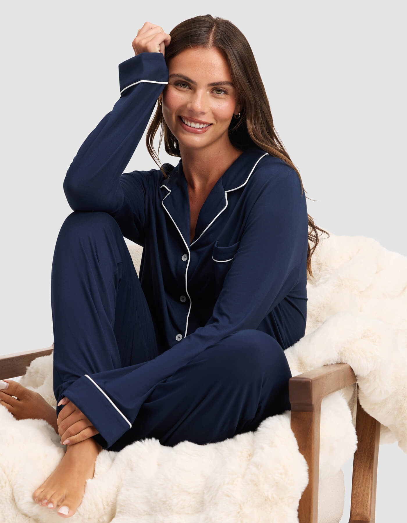 A woman with long brown hair smiles barefoot on a white faux fur chair, wearing the Cozy Earth Women's Bamboo Stretch-Knit Long Sleeve Pajama Top in navy blue with white piping, set against a light gray background. 