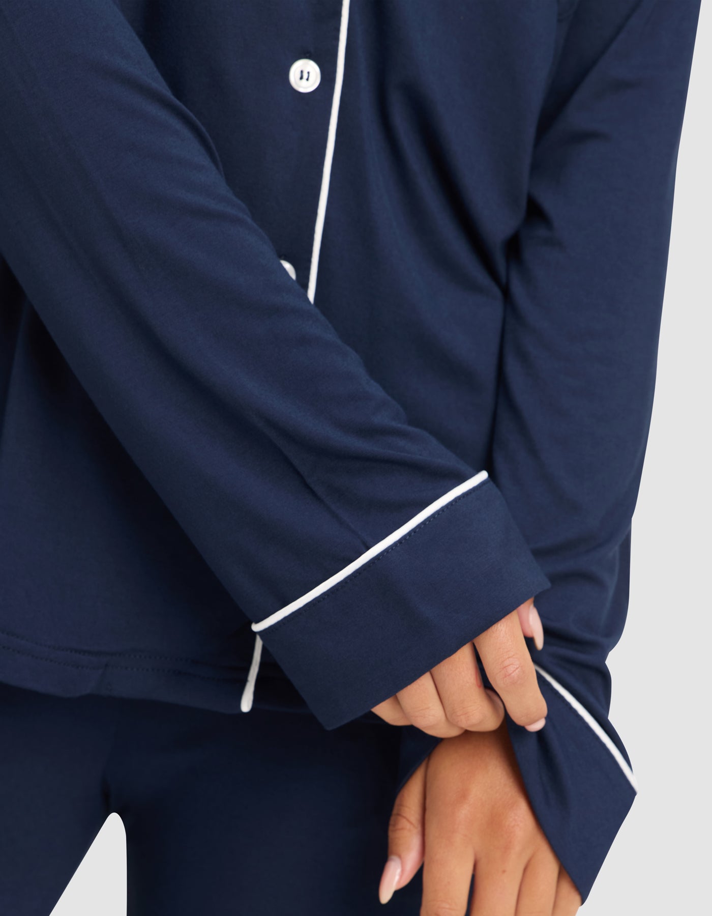A woman models the HIDE Women's Bamboo Stretch-Knit Long Sleeve Pajama Top in navy with white piping, shown from the shoulders down as she pulls at her sleeve. Only her lower body and hands are partially visible. 