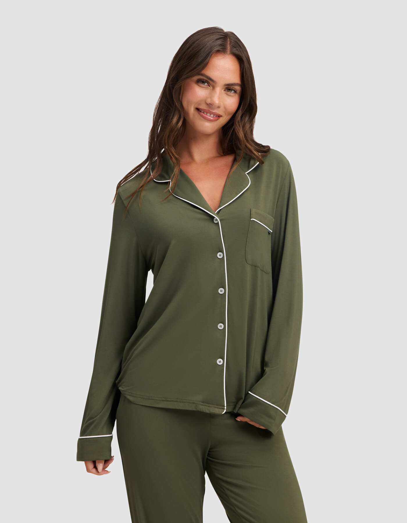 A woman with long brown hair smiles in a Cozy Earth Women's Bamboo Stretch-Knit Long Sleeve Pajama Top, olive green with white piping and a chest pocket, paired with matching pants against a plain light gray background.