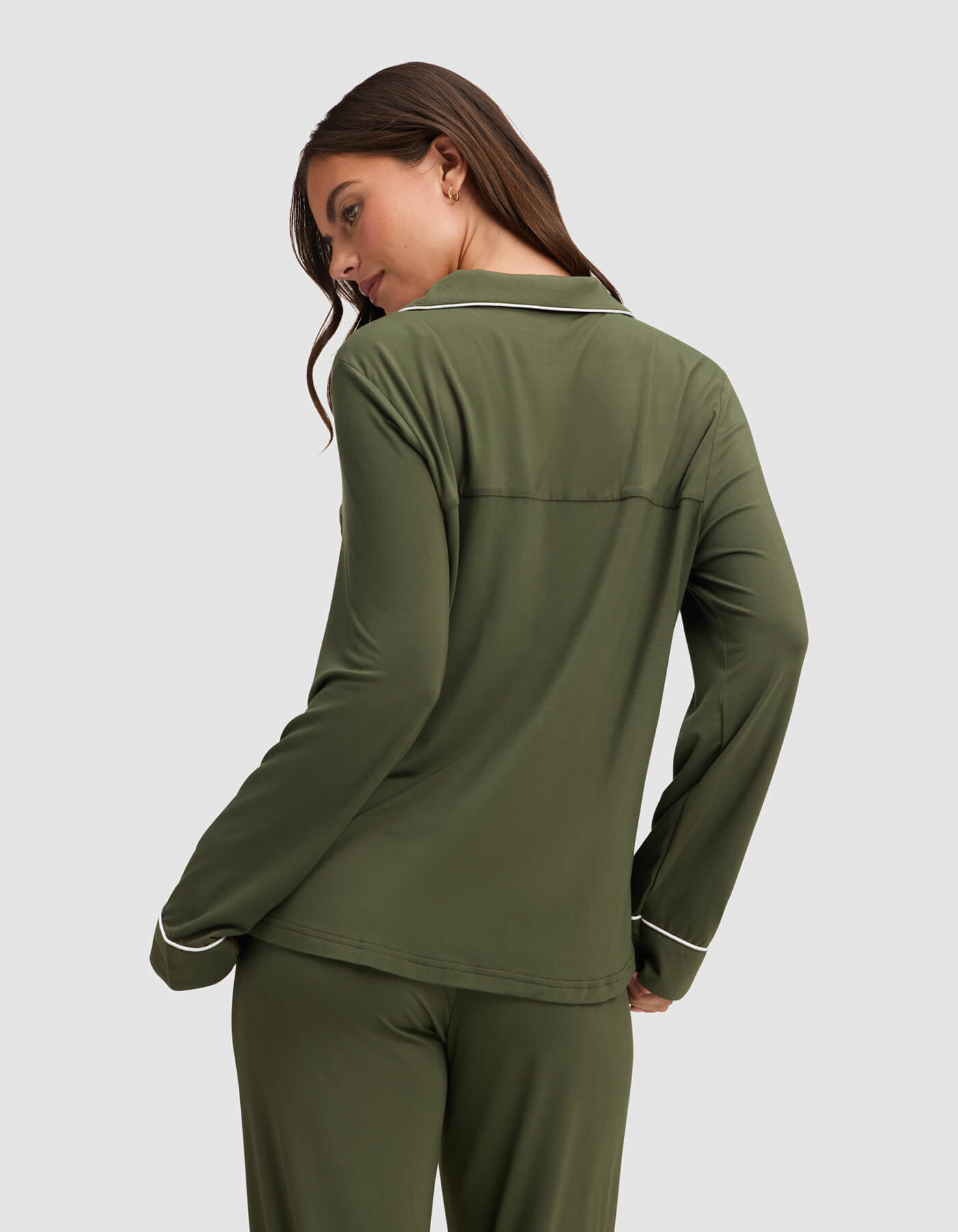 A woman with long brown hair wears the Cozy Earth Women's Bamboo Stretch-Knit Long Sleeve Pajama Top in olive green with white piping, standing slightly turned to the side and looking over her shoulder against a light gray background.