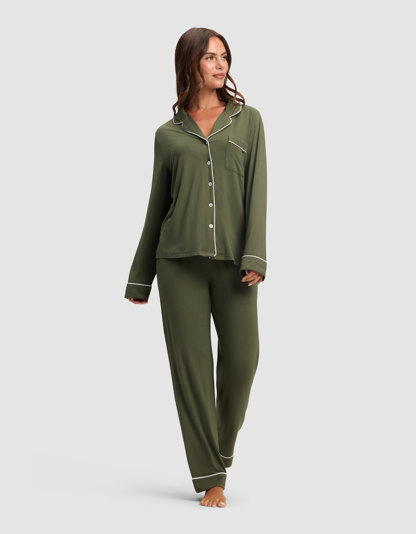 A woman with long brown hair smiles and looks to the side, barefoot against a plain light background, wearing the HIDE Women's Bamboo Stretch-Knit Long Sleeve Pajama Top in olive green with white piping and matching pants.