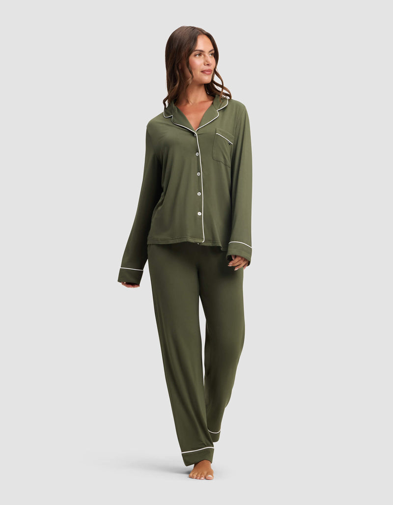 A woman stands barefoot against a light gray background, wearing Cozy Earth's Women's Bamboo Stretch-Knit Long Sleeve Pajama Top in olive green with white piping, paired with matching pants.