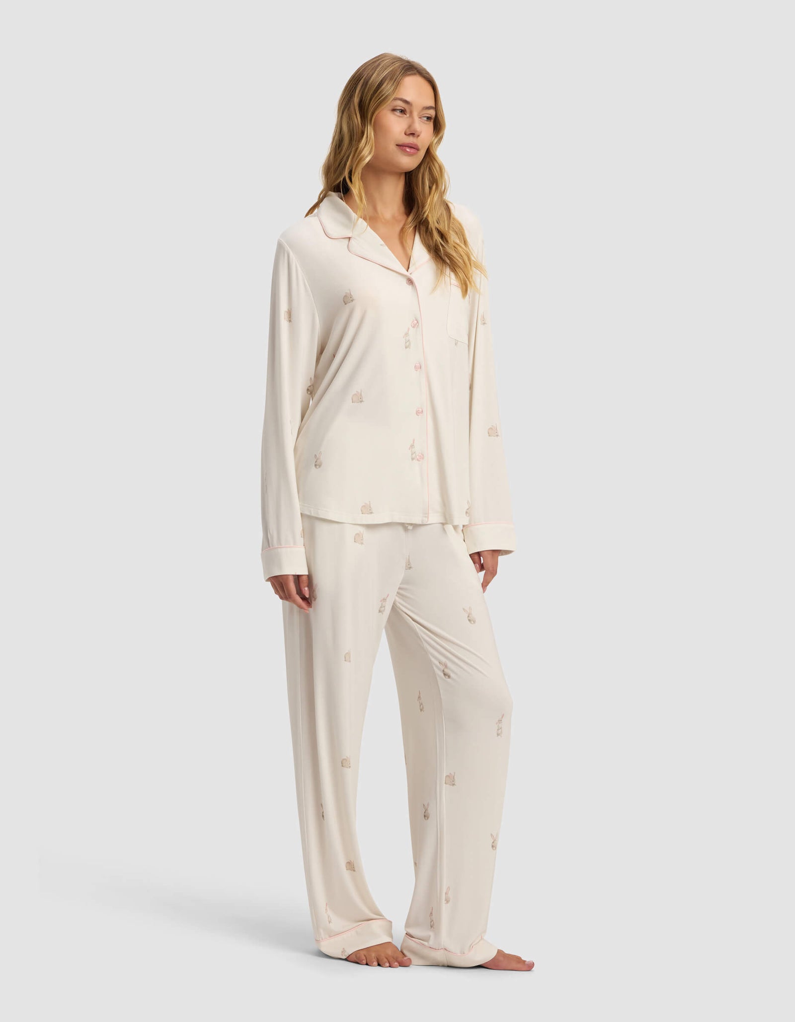 A woman stands barefoot in Cozy Earth's Women's Bamboo Stretch-Knit Long Sleeve Pajama Set, featuring a cream-colored button-up top and loose pants with a subtle small print pattern.