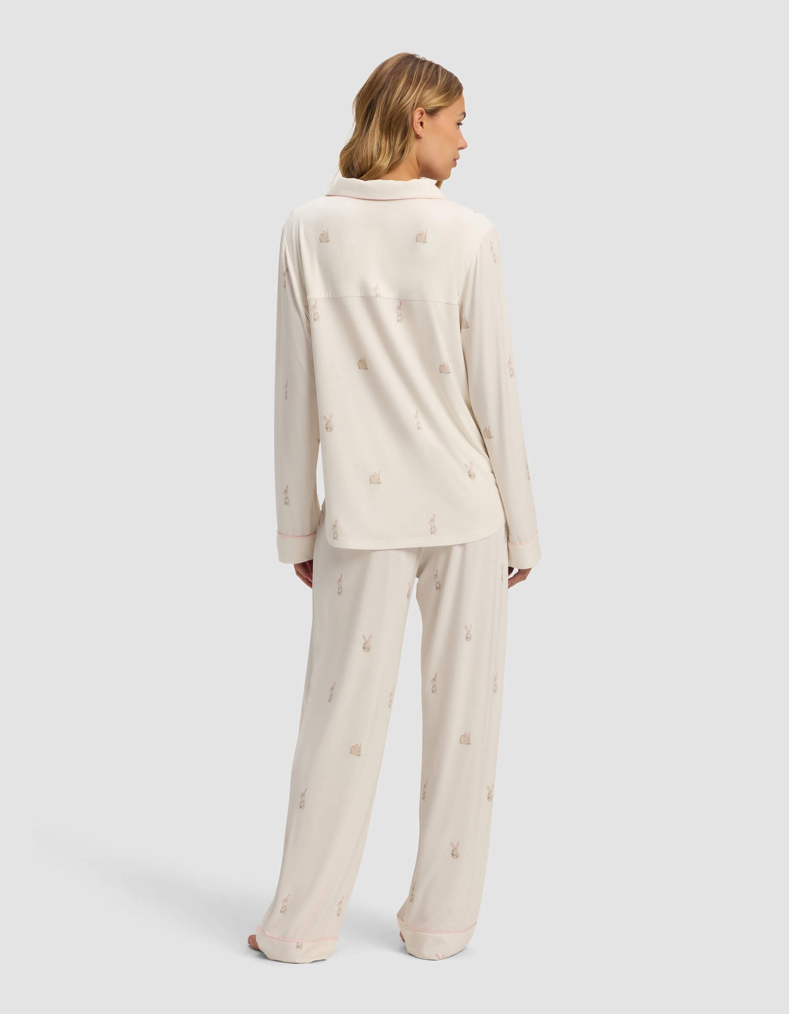 A woman with shoulder-length hair and bare feet faces away, wearing the Cozy Earth Women's Bamboo Stretch-Knit Long Sleeve Pajama Set in cream with a subtle small print. The set features a long-sleeved top and loose-fitting pants against a light background.