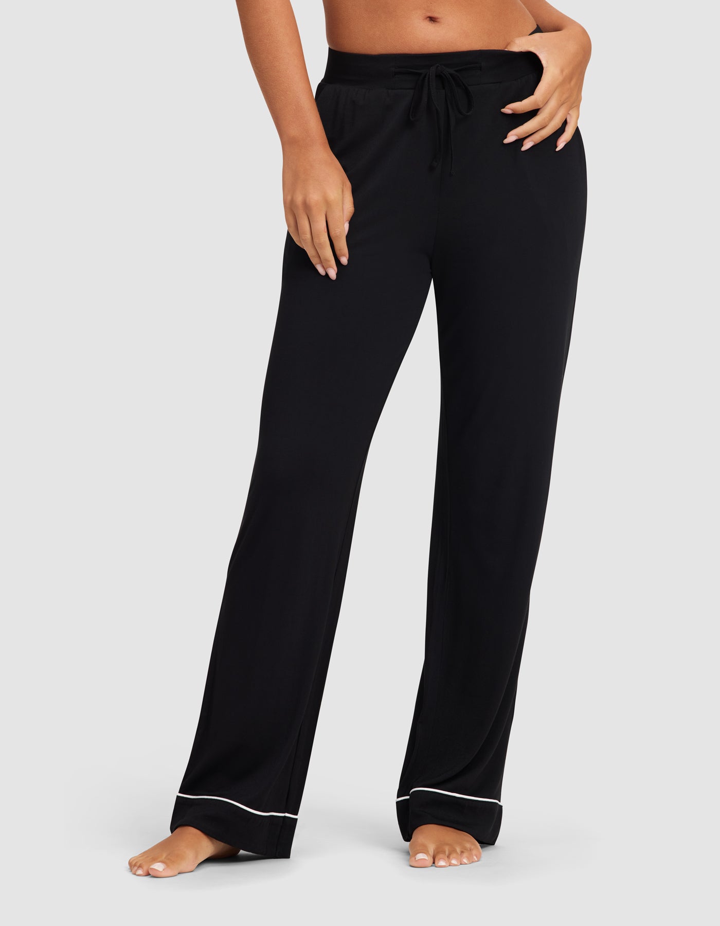 A woman models the HIDE Women's Bamboo Stretch-Knit Pajama Pant in black, featuring a drawstring waist and white piping at the hem, standing barefoot against a light gray background.