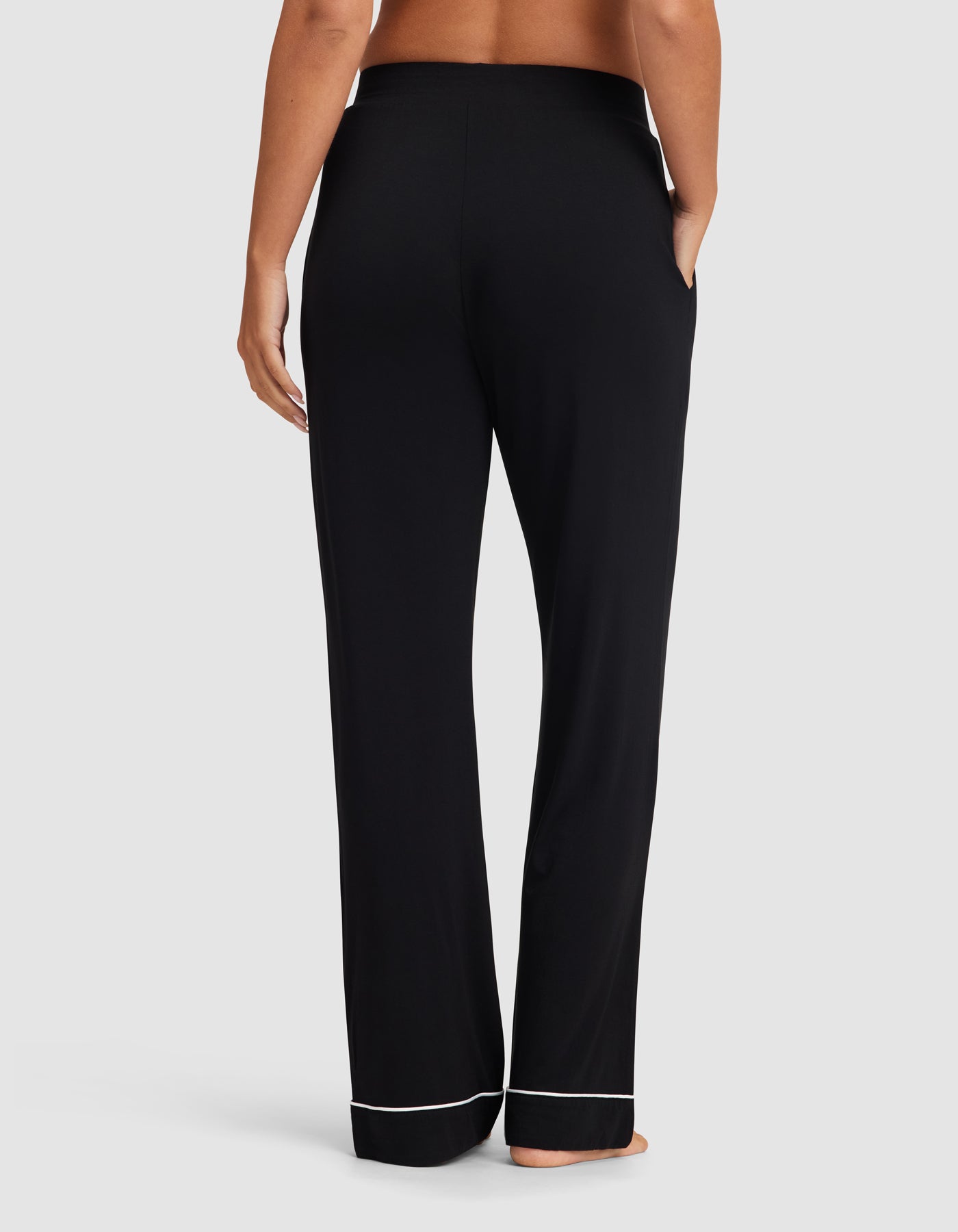 A woman stands barefoot, shown from the back, wearing HIDE Women's Bamboo Stretch-Knit Pajama Pant in black with pockets and white hem trim against a plain light background.