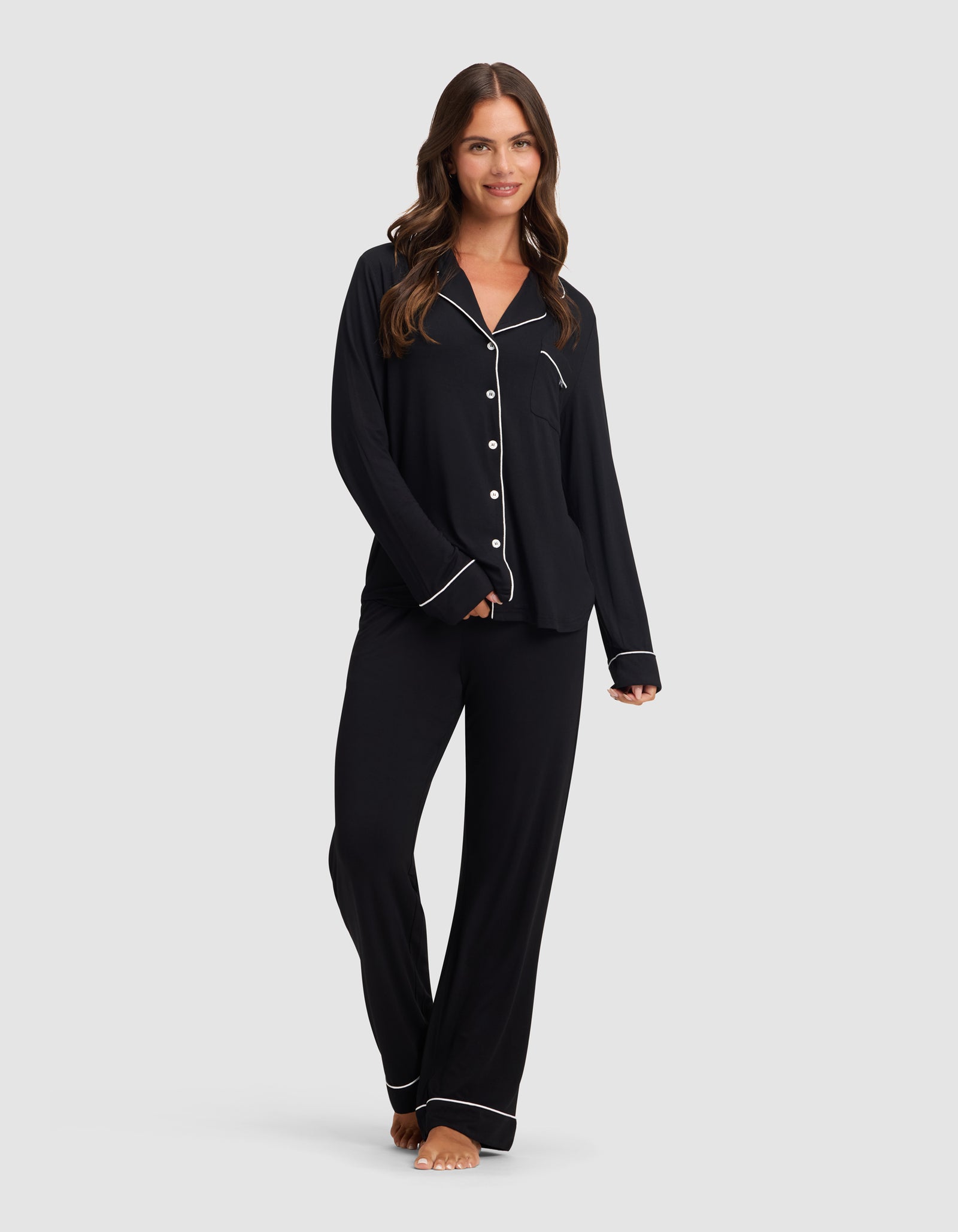 A woman with long brown hair, barefoot and smiling, models the HIDE Women's Bamboo Stretch-Knit Pajama Pant in black with white piping, paired with a matching button-up top, against a plain light gray background.