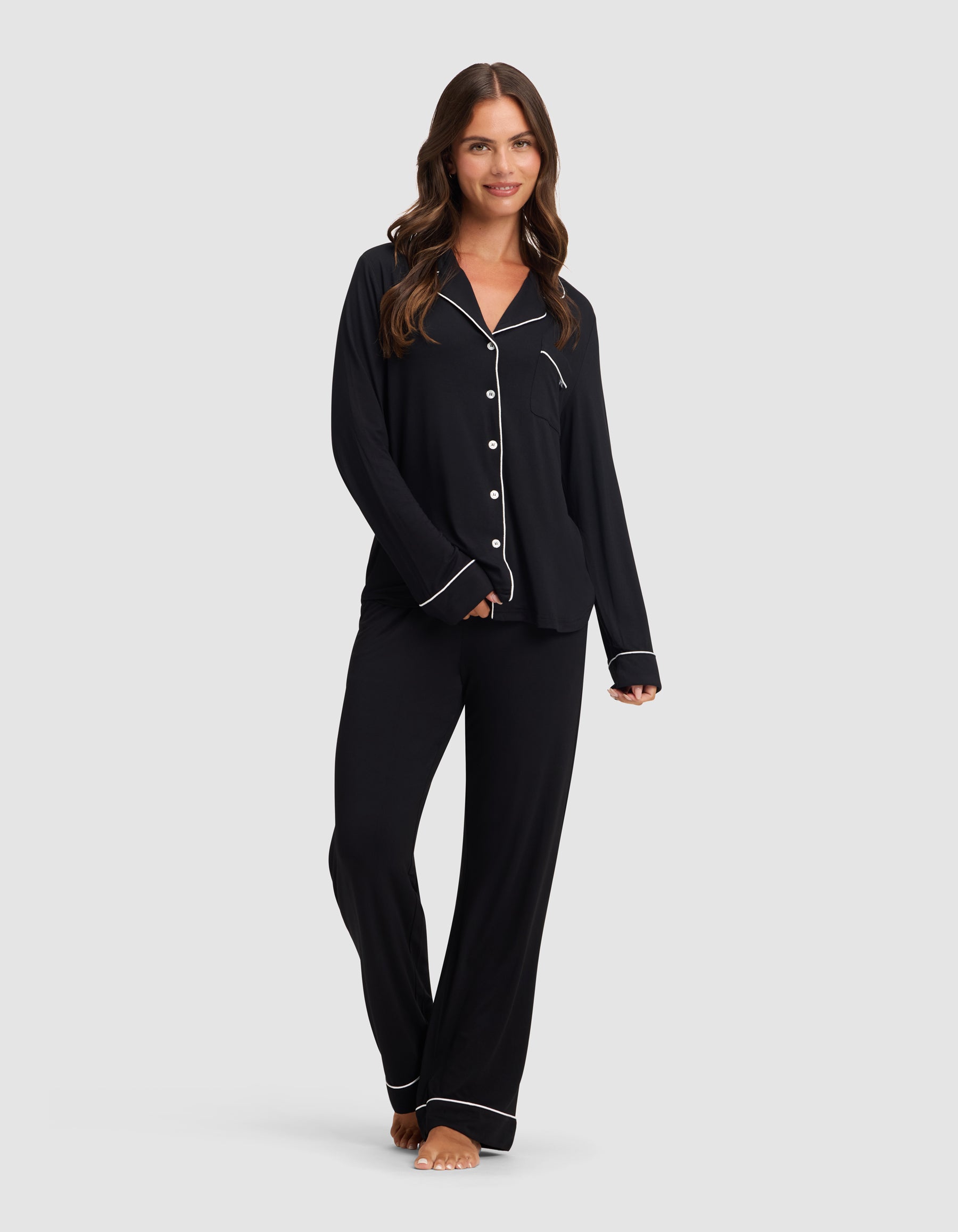 A woman with long brown hair, barefoot and smiling, models the HIDE Women's Bamboo Stretch-Knit Pajama Pant in black with white piping, paired with a matching button-up top, against a plain light gray background.