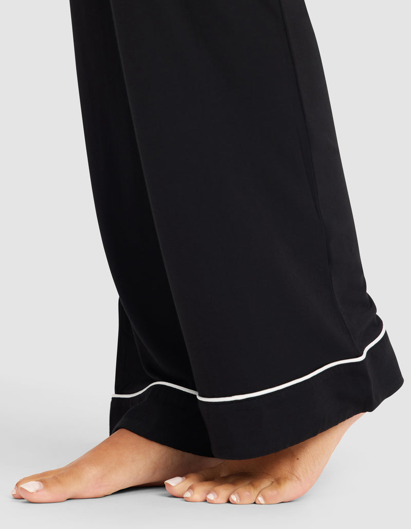 A close-up of bare feet on the floor in HIDE Women's Bamboo Stretch-Knit Pajama Pant, featuring black fabric with white piping on the hem, set against a plain light background.