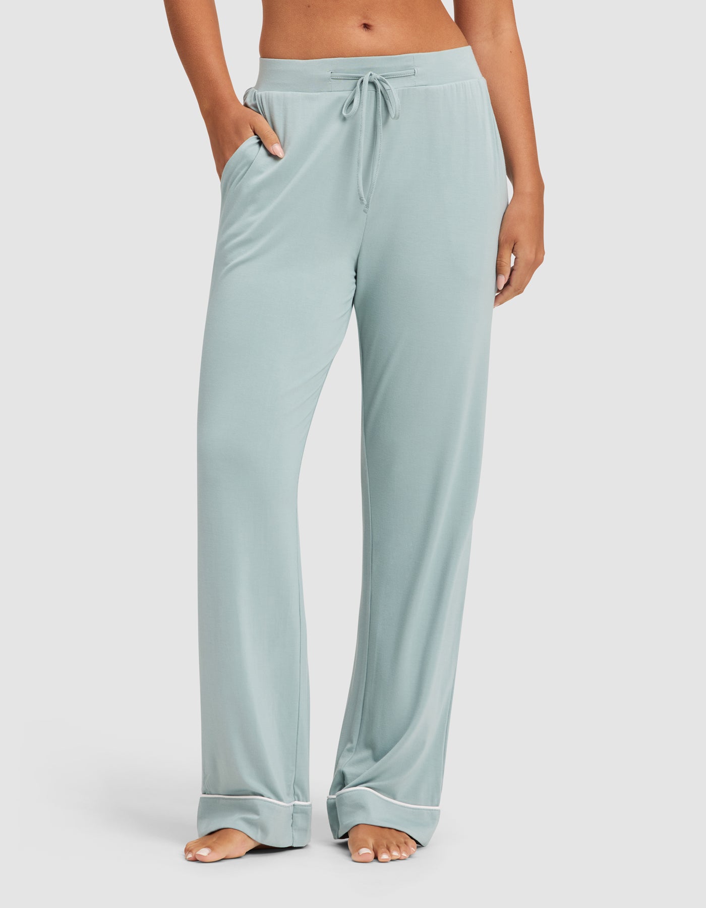 A person stands barefoot with hands in the pockets of HIDE Women's Bamboo Stretch-Knit Pajama Pant in light blue. The relaxed-fit, drawstring pants have side pockets, and the photo is cropped at their waist and ankles. 