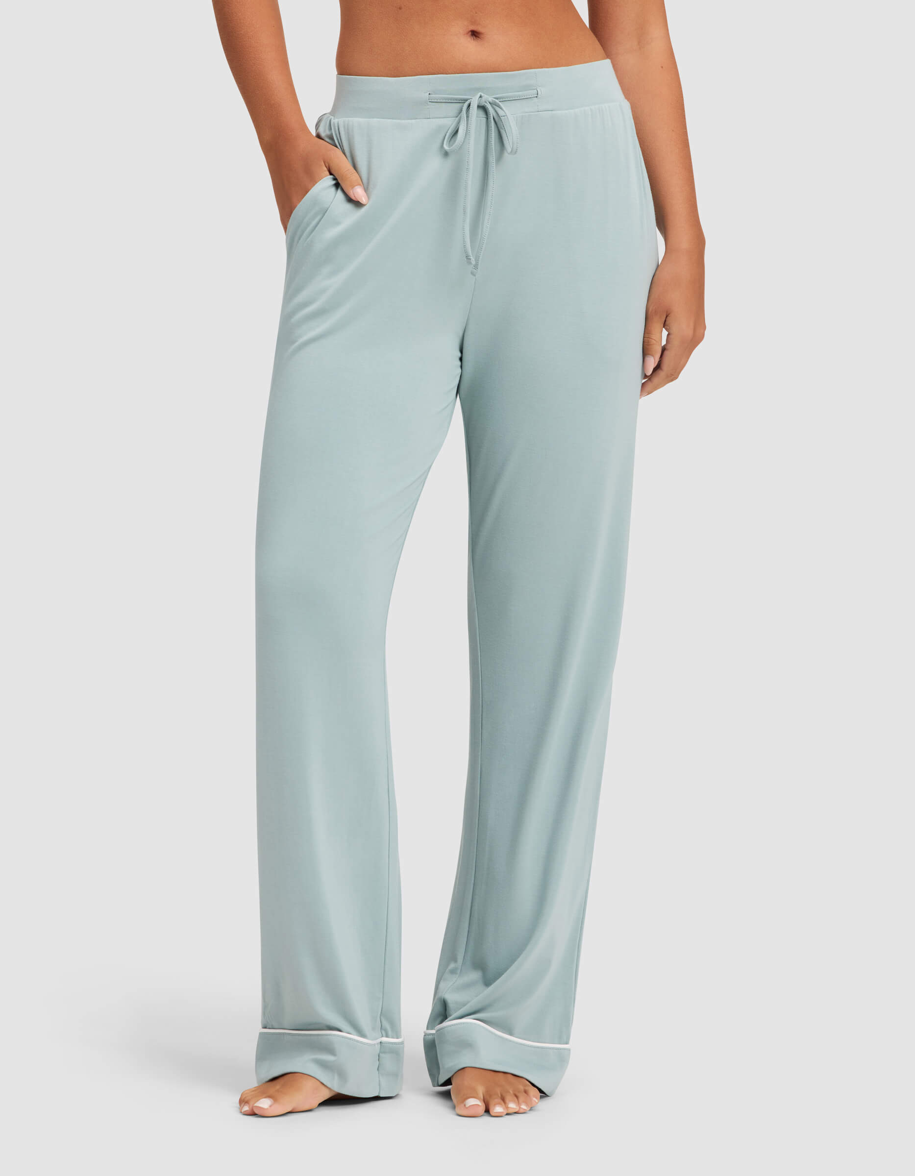 A person stands barefoot on a white background wearing Cozy Earth's Women's Bamboo Stretch-Knit Pajama Pant in light blue. The loose-fitting pants feature a drawstring and pockets, with the image cropped above the waist.|Color:Brook