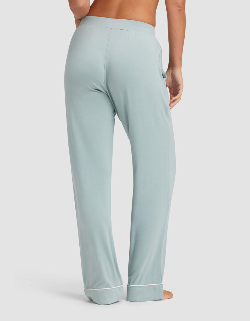 A woman stands barefoot, facing away, in Cozy Earth's Women's Bamboo Stretch-Knit Pajama Pant—light blue with white-trimmed cuffs, relaxed fit, and elastic waistband—against a plain light background.