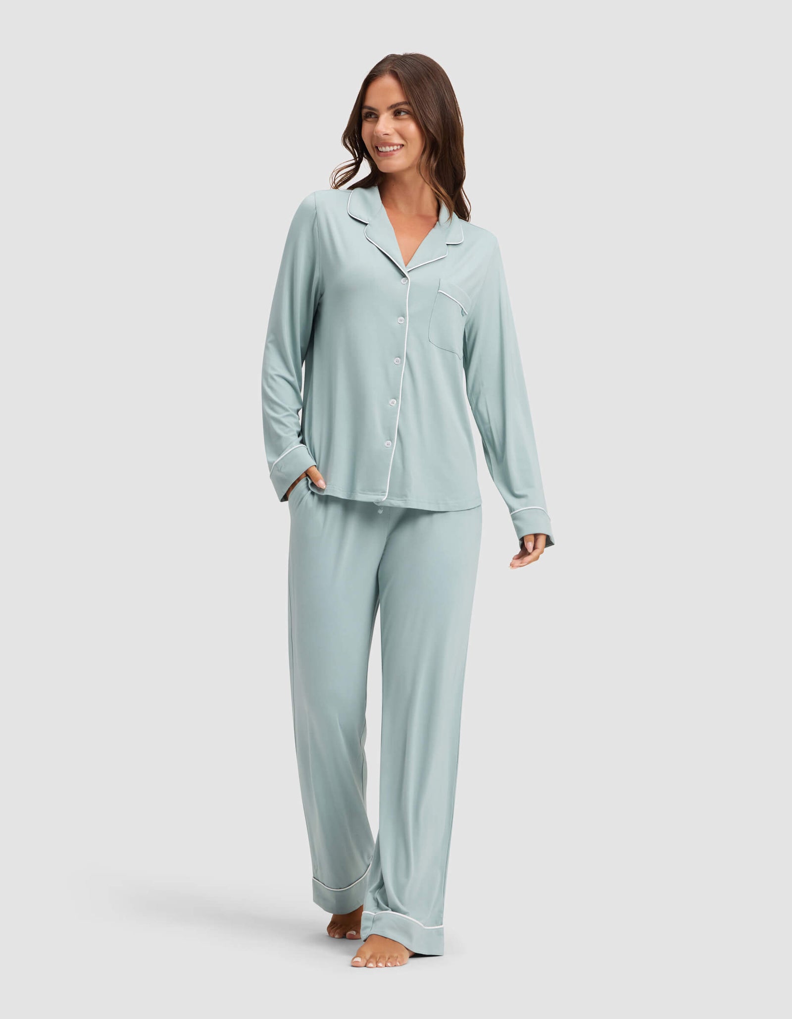 A woman smiles while wearing Cozy Earth Women's Bamboo Stretch-Knit Pajama Pant in light blue with a matching long-sleeve button-up top, barefoot against a plain light background. 