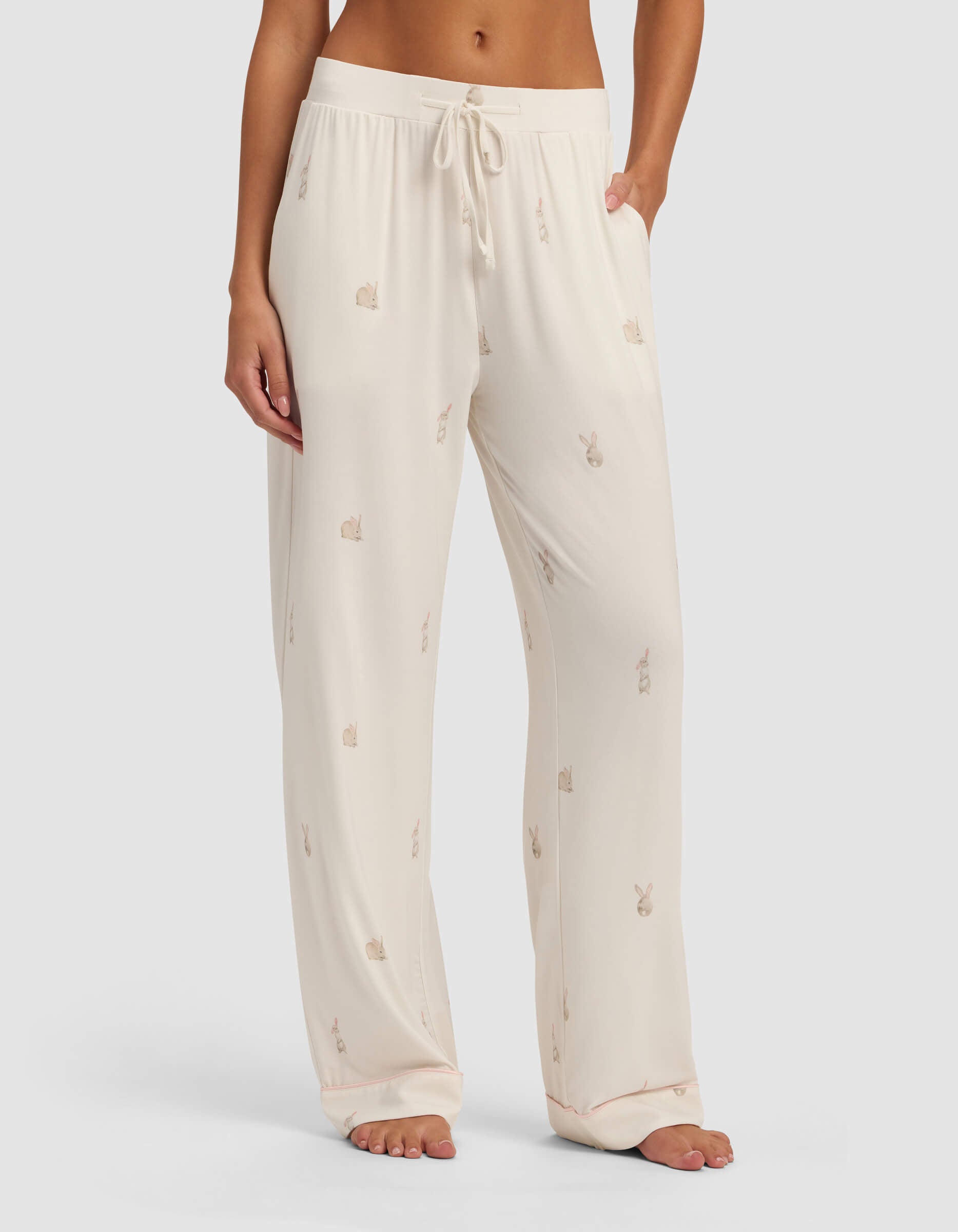 A person stands barefoot against a plain background wearing Cozy Earth Women's Bamboo Stretch-Knit Pajama Pant, featuring a drawstring waist and small bunny designs in a loose, cream-colored style.