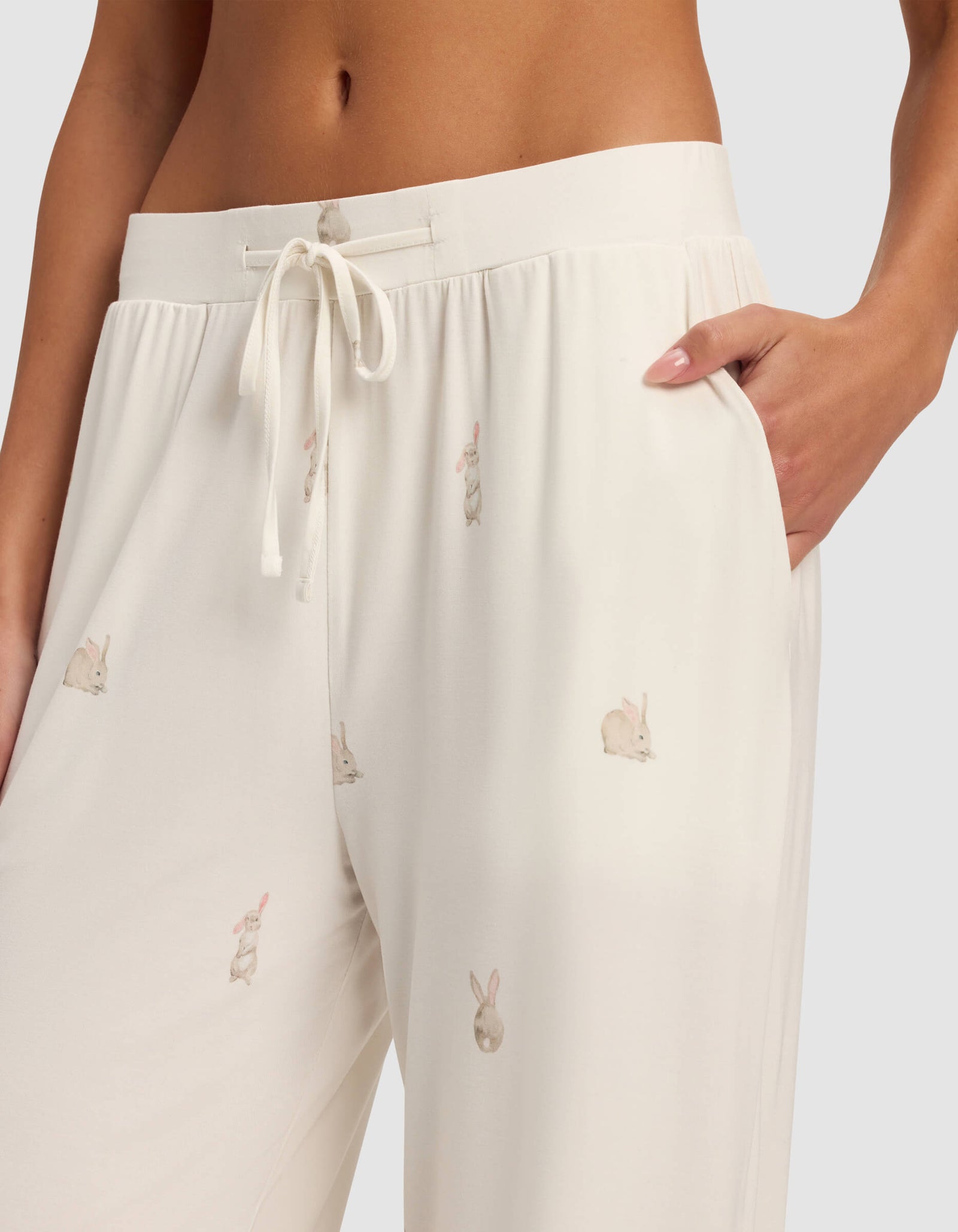 A close-up of a person wearing Cozy Earth's Women's Bamboo Stretch-Knit Pajama Pant in white, with a drawstring, pockets, and light brown bunny pattern. One hand is placed in the pocket.