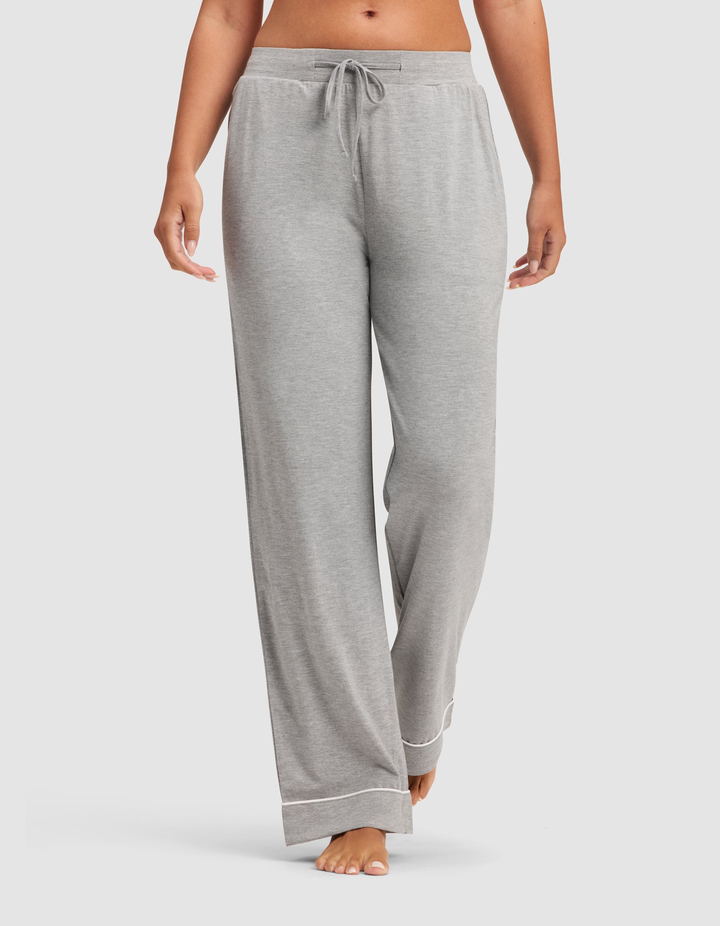 A woman models the HIDE Women's Bamboo Stretch-Knit Pajama Pant in light grey, featuring a drawstring waistband and white hem trim, standing barefoot against a plain white background. 