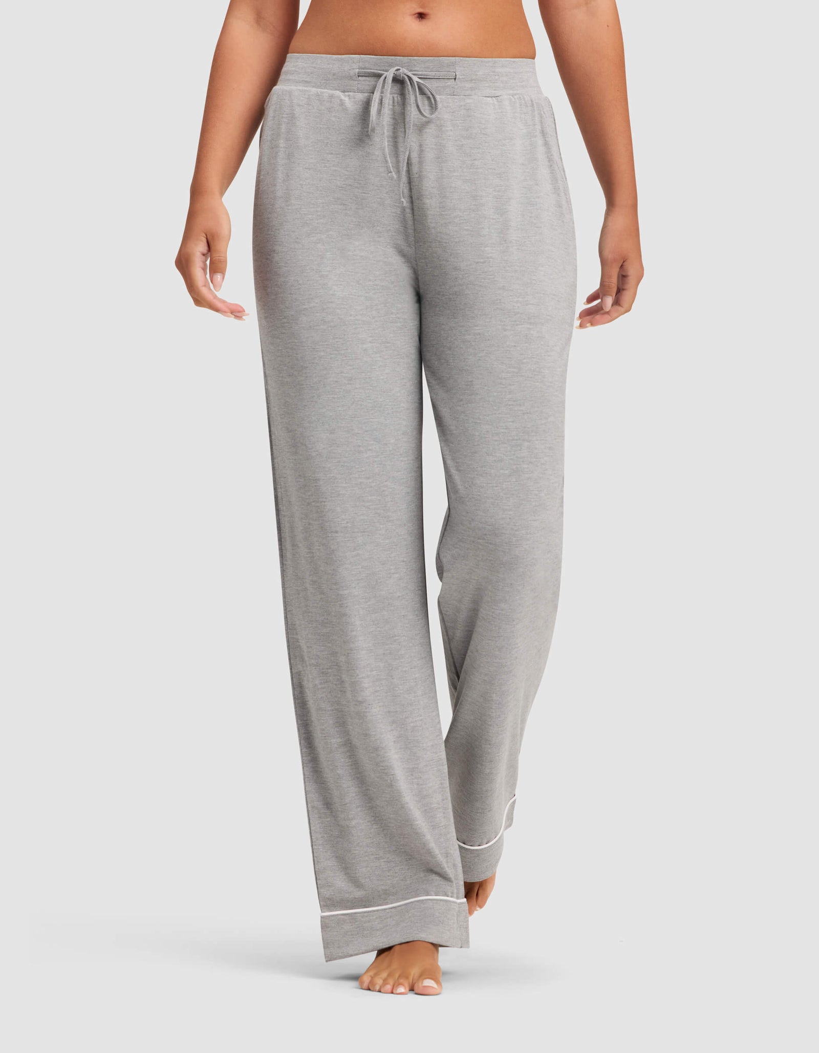 A woman wears Cozy Earth Women's Bamboo Stretch-Knit Pajama Pants in light gray, featuring a drawstring waist and white trim at the cuffs, standing barefoot against a plain background.