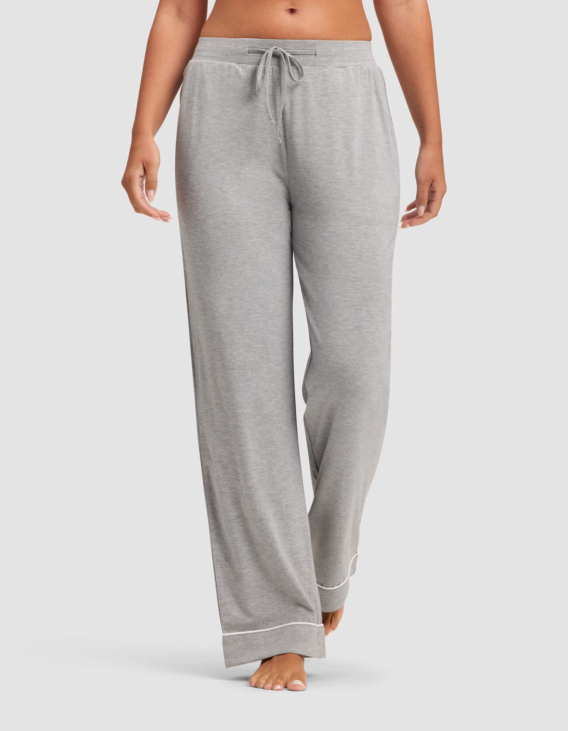 A woman wears Cozy Earth Women's Bamboo Stretch-Knit Pajama Pants in light gray, featuring a drawstring waist and white trim at the cuffs, standing barefoot against a plain background.|Color:Grey