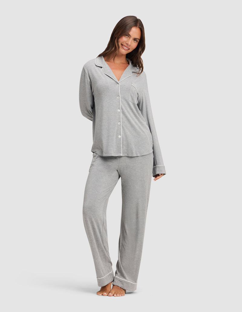 A woman stands barefoot, smiling, and wearing HIDE’s Women's Bamboo Stretch-Knit Pajama Pant in light gray with a matching button-up long-sleeve top, against a plain white background.