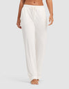 A model wears HIDE Women's Bamboo Stretch-Knit Pajama Pants in white, featuring a loose fit and drawstring waist, barefoot against a plain light gray background. Only the lower torso and legs are shown.|Color:Ivory