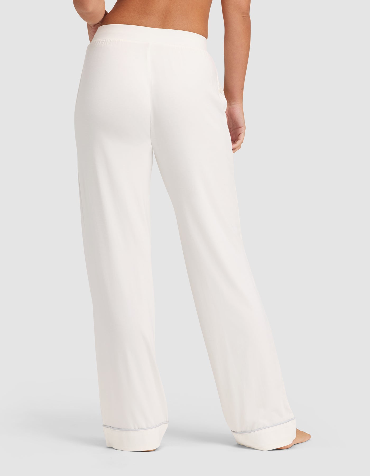 A person is shown from the waist down wearing HIDE Women's Bamboo Stretch-Knit Pajama Pant in white, featuring a wide waistband, relaxed straight-leg fit, and lightly pooled hems against a plain light-colored background. 