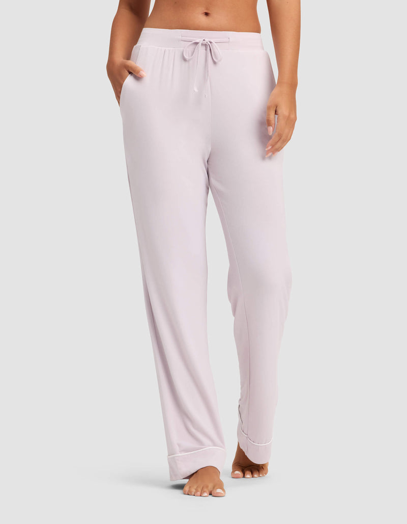 A woman models the Cozy Earth Women's Bamboo Stretch-Knit Pajama Pant in light pink, featuring a drawstring waist and pockets, while standing barefoot against a plain light background.