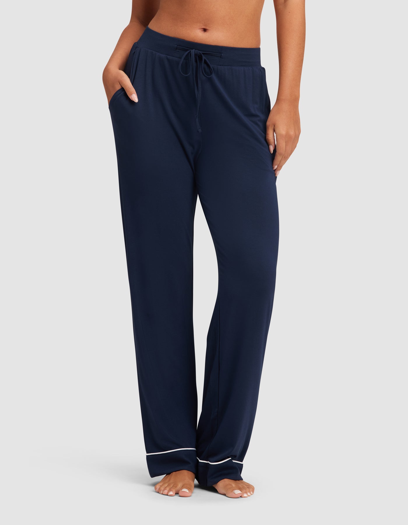 A woman wears HIDE Women's Bamboo Stretch-Knit Pajama Pant in navy, featuring white piping near the ankles, standing barefoot with hands in pockets against a plain light background. 