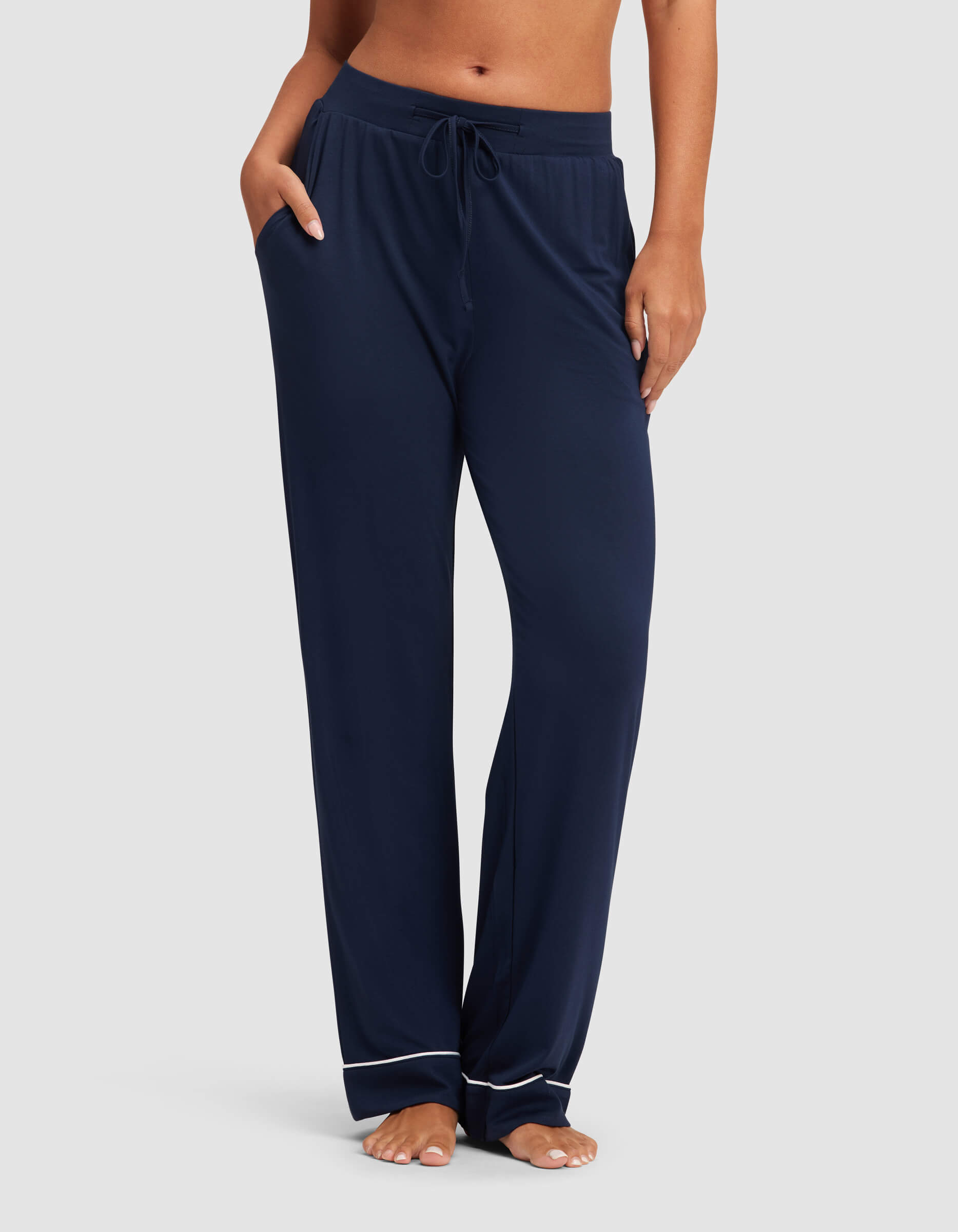 A woman stands barefoot against a plain background wearing Cozy Earth's Women's Bamboo Stretch-Knit Pajama Pant, featuring a loose fit, drawstring waist, and white piping near the hems.