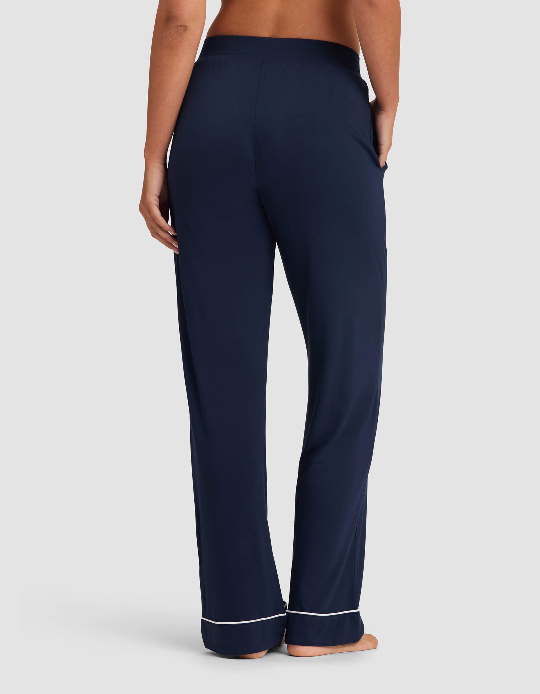 A woman stands barefoot facing away, wearing Cozy Earth Women's Bamboo Stretch-Knit Pajama Pant in navy with white piping at the cuffs and one hand in a pocket against a plain light gray background.