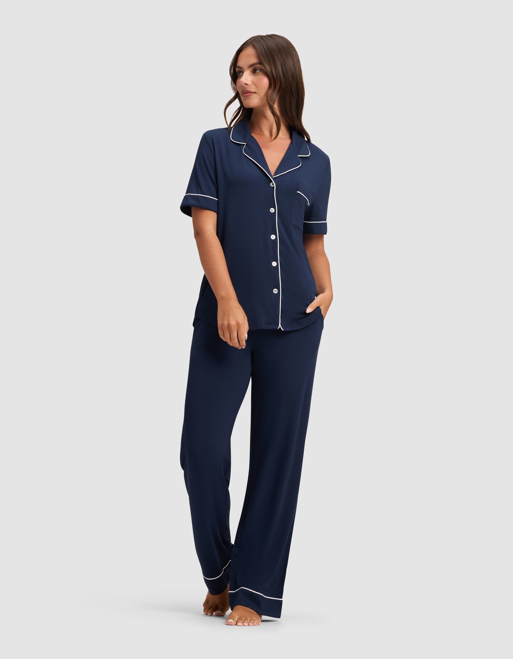 A woman stands barefoot, looking to the side, wearing HIDE's Women's Bamboo Stretch-Knit Pajama Pant in navy blue with white piping. The pajama top features short sleeves, a collar, and buttons; the pants are loose-fitting. |Color:Navy