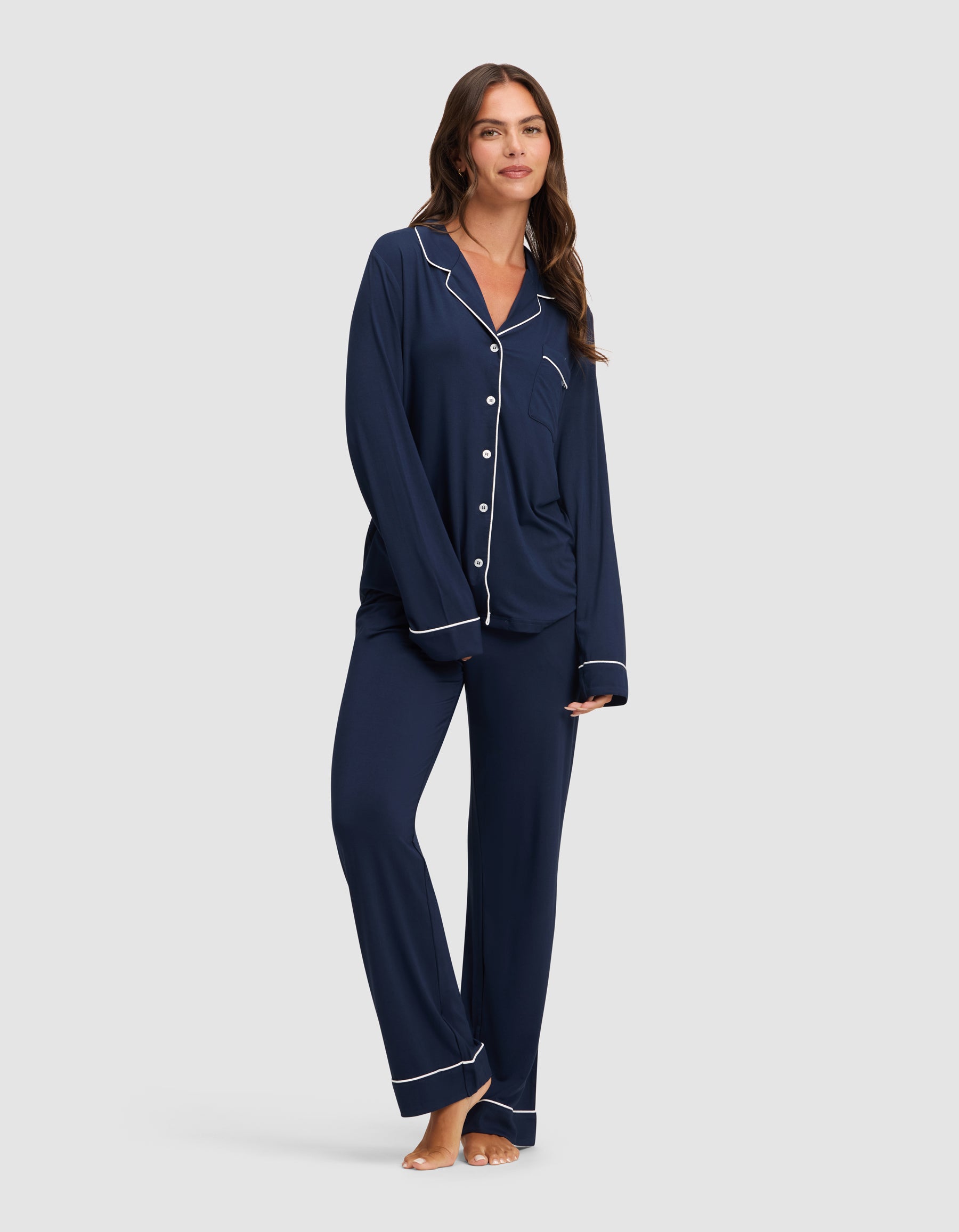 A woman stands barefoot against a light gray background, wearing HIDE's Women's Bamboo Stretch-Knit Pajama Pant in navy blue with white piping and a matching button-up long-sleeve top. 