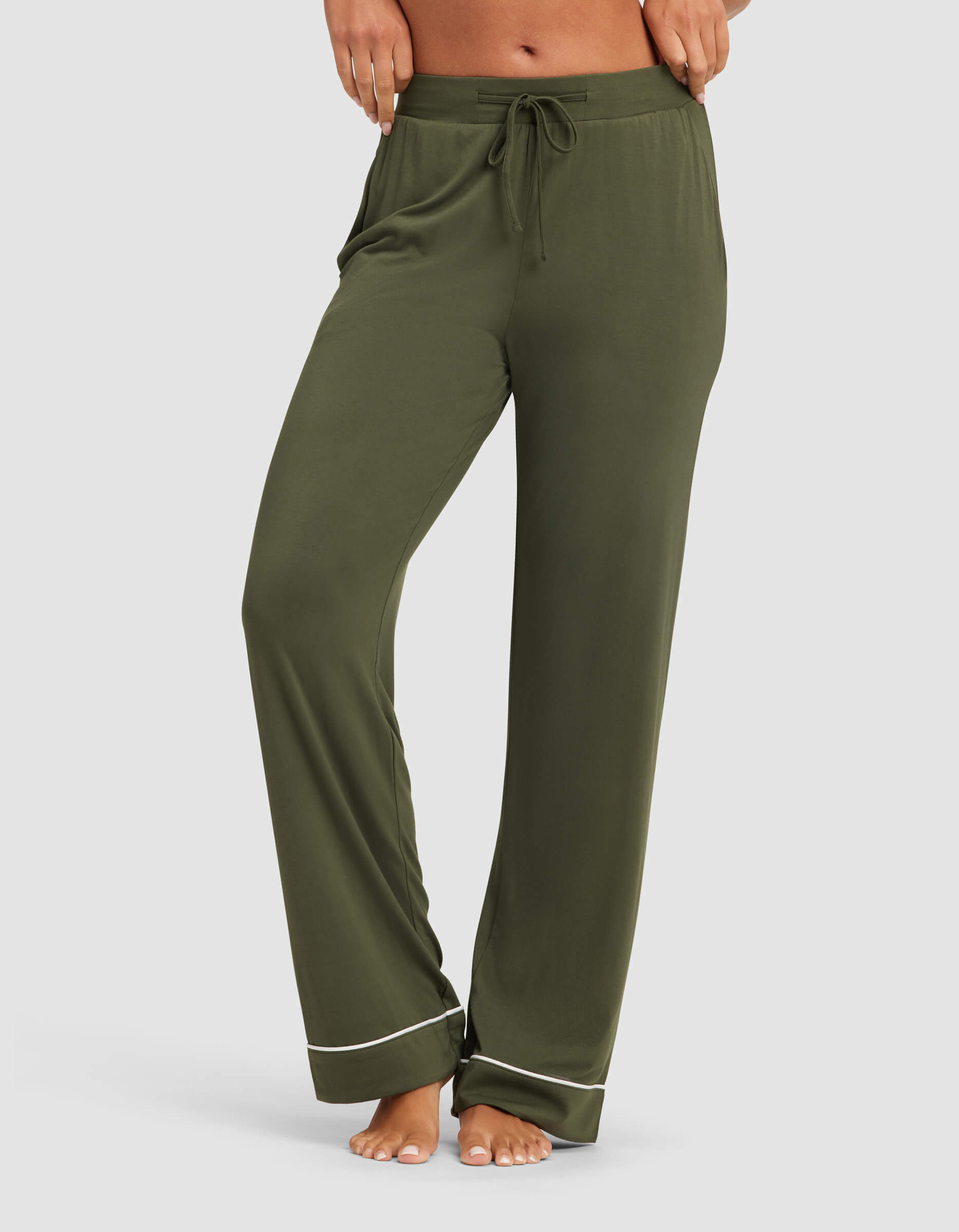 A model showcases the lower half in Cozy Earth Women's Bamboo Stretch-Knit Pajama Pant, featuring a loose olive green fit, drawstring waist, and white hem piping. The model is barefoot.