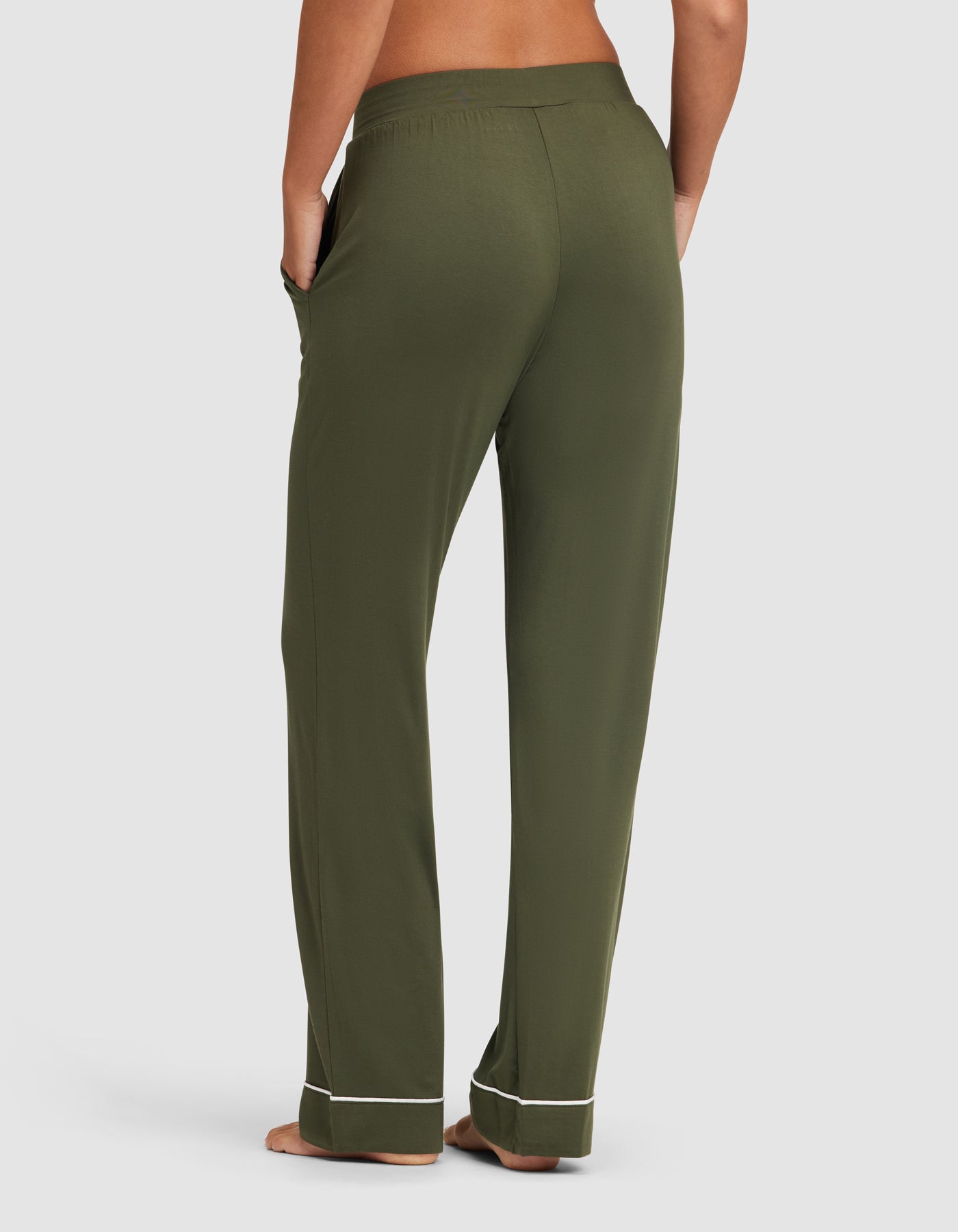 A person stands barefoot, facing away from the camera, wearing HIDE Women's Bamboo Stretch-Knit Pajama Pant in olive green with white piping at the cuffs. Their hands are in the pockets against a plain white background. 