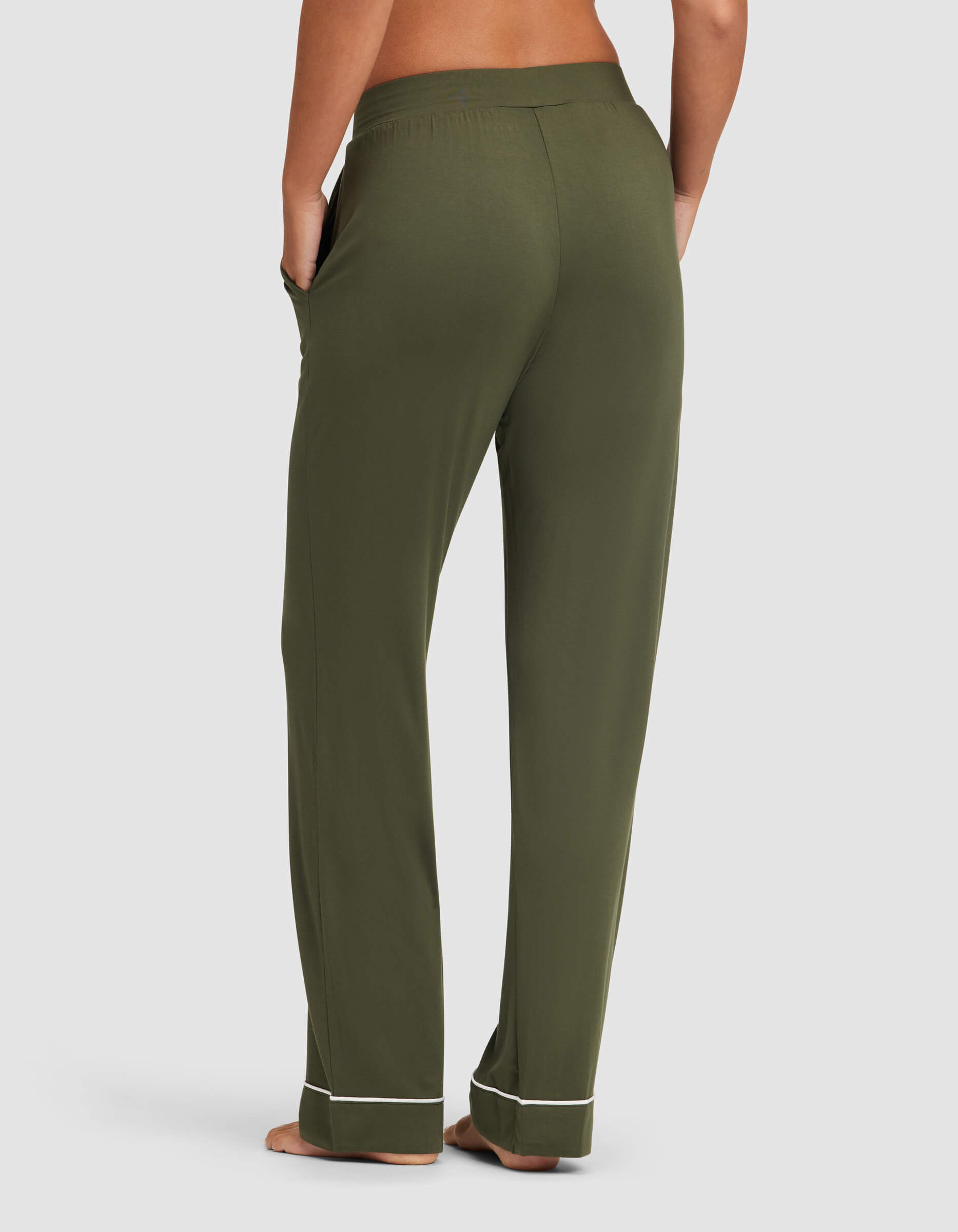 A person stands barefoot facing away, wearing Cozy Earth's Women's Bamboo Stretch-Knit Pajama Pant in olive green with white piping near the cuffs and hands in pockets, set against a plain light background.