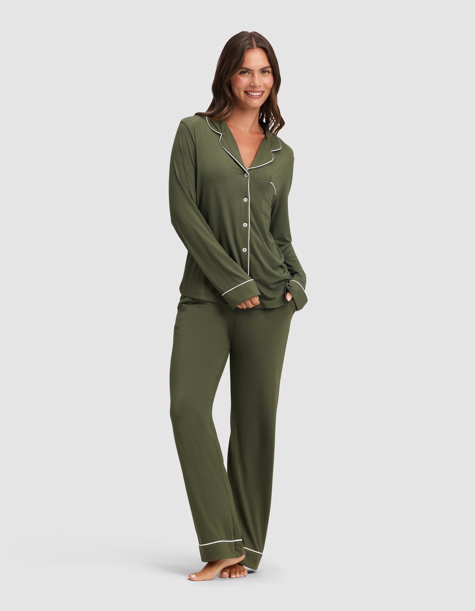 A woman smiles, barefoot, in HIDE's Women's Bamboo Stretch-Knit Pajama Pant—an olive green set with white piping and a button-up long-sleeve top—posing with one hand in her pocket against a plain background. 