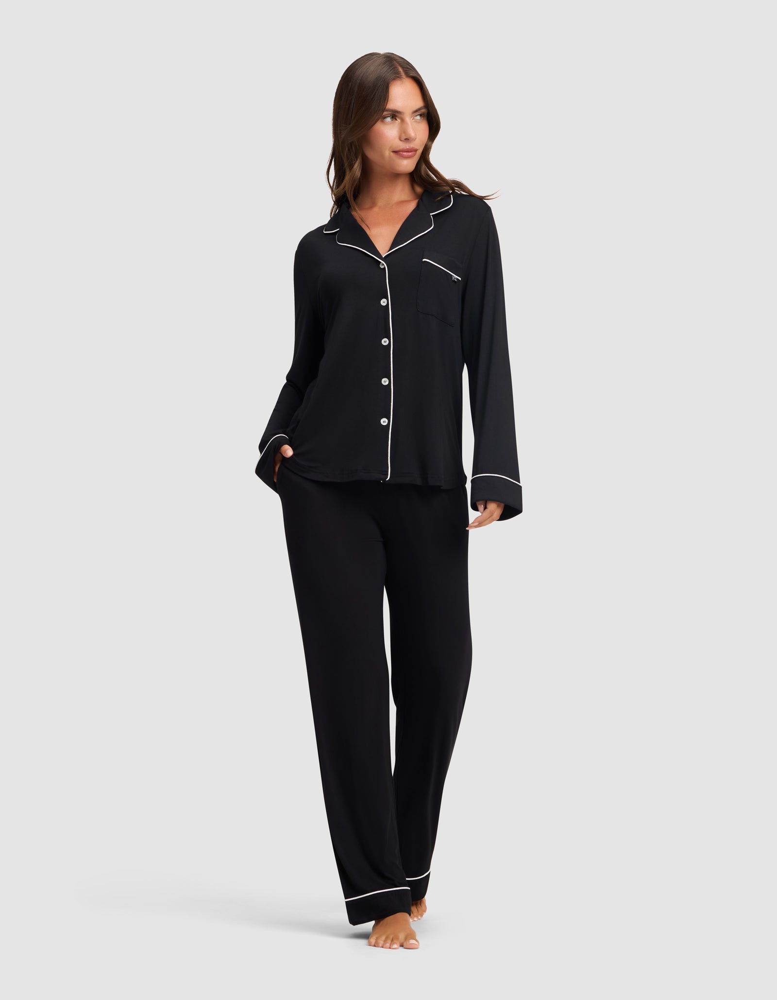 A woman stands barefoot against a light gray background, wearing Cozy Earth's Women's Bamboo Stretch-Knit Long Sleeve Pajama Set in black with white piping, featuring a button-up top and matching pants. 