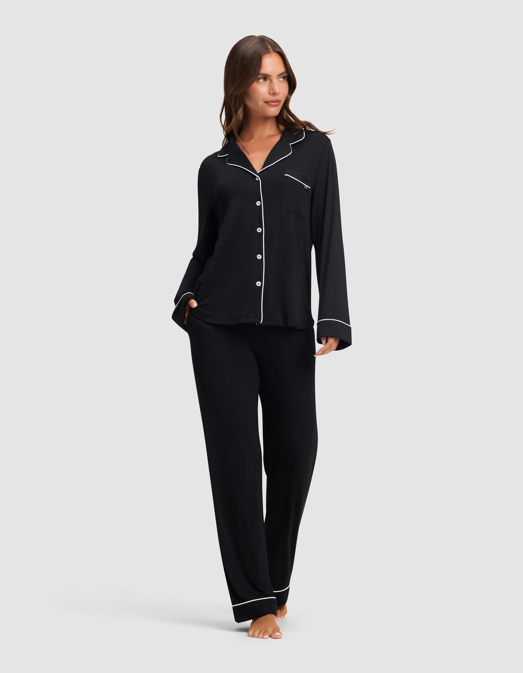A woman stands barefoot against a light gray background, wearing Cozy Earth's Women's Bamboo Stretch-Knit Long Sleeve Pajama Set in black with white piping, featuring a button-up top and matching pants. 