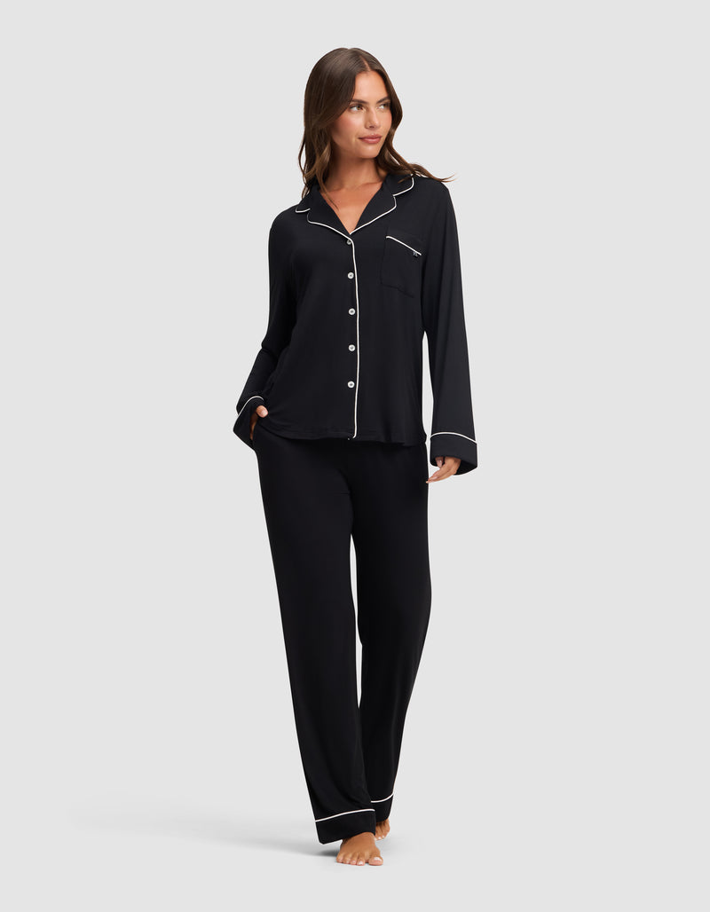 A woman stands barefoot against a light gray background, wearing Cozy Earth's Women's Bamboo Stretch-Knit Long Sleeve Pajama Set in black with white piping, featuring a button-up top and matching pants.