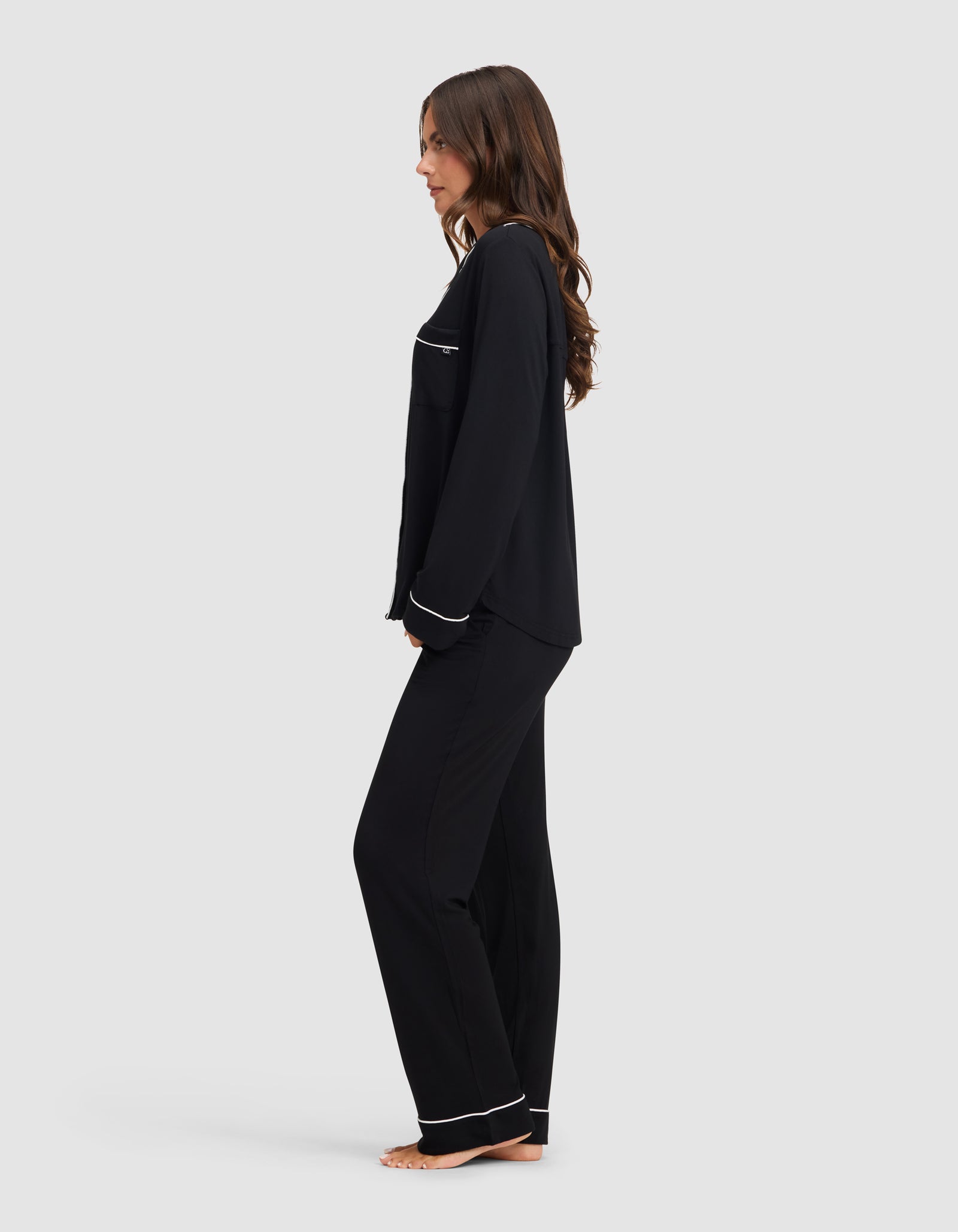 A woman stands barefoot in profile against a light gray background, wearing the Cozy Earth Women's Bamboo Stretch-Knit Long Sleeve Pajama Set in black with white trim. 