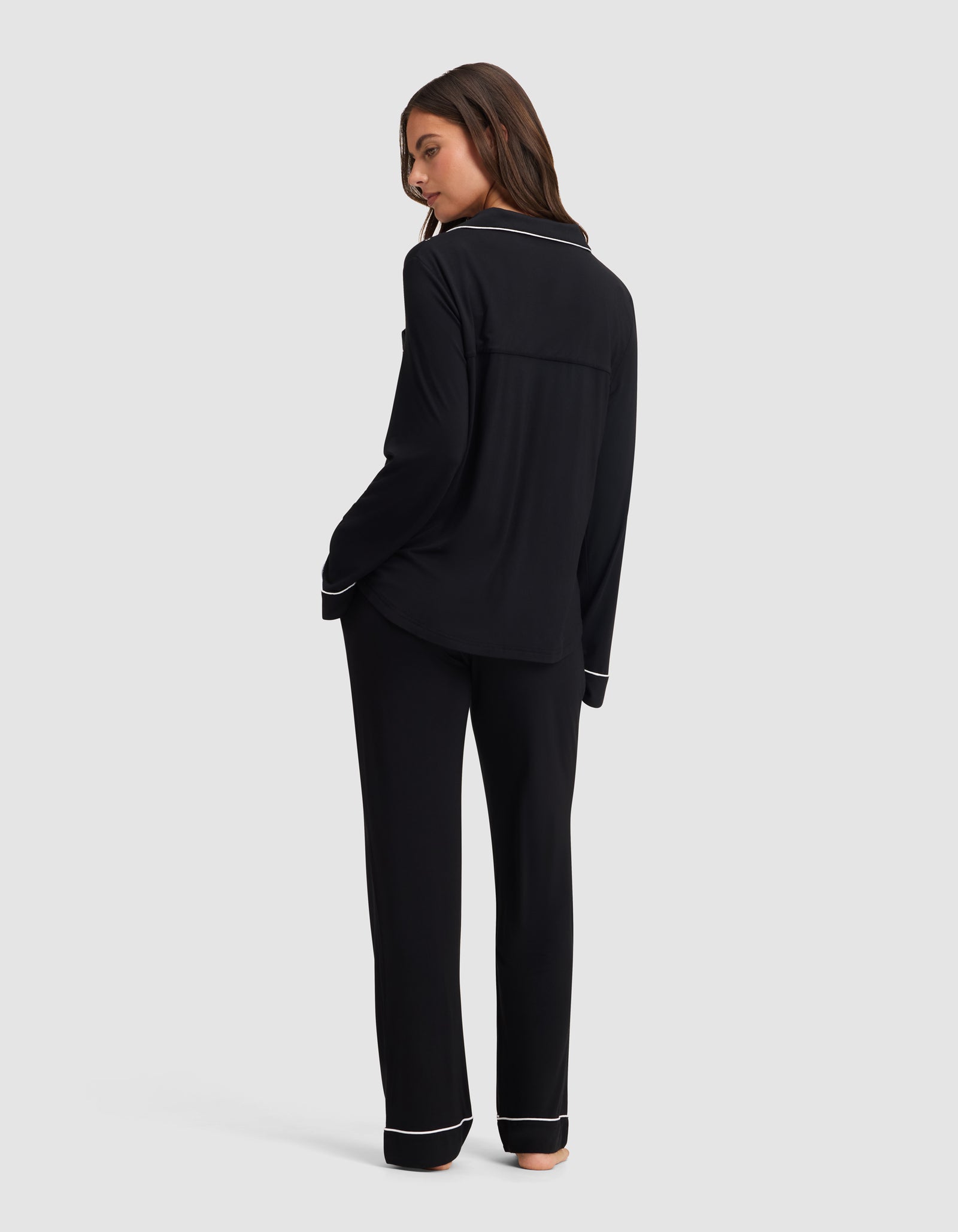 A woman stands barefoot, turned slightly away, wearing the Cozy Earth Women's Bamboo Stretch-Knit Long Sleeve Pajama Set in black with white piping, against a plain light gray background. 