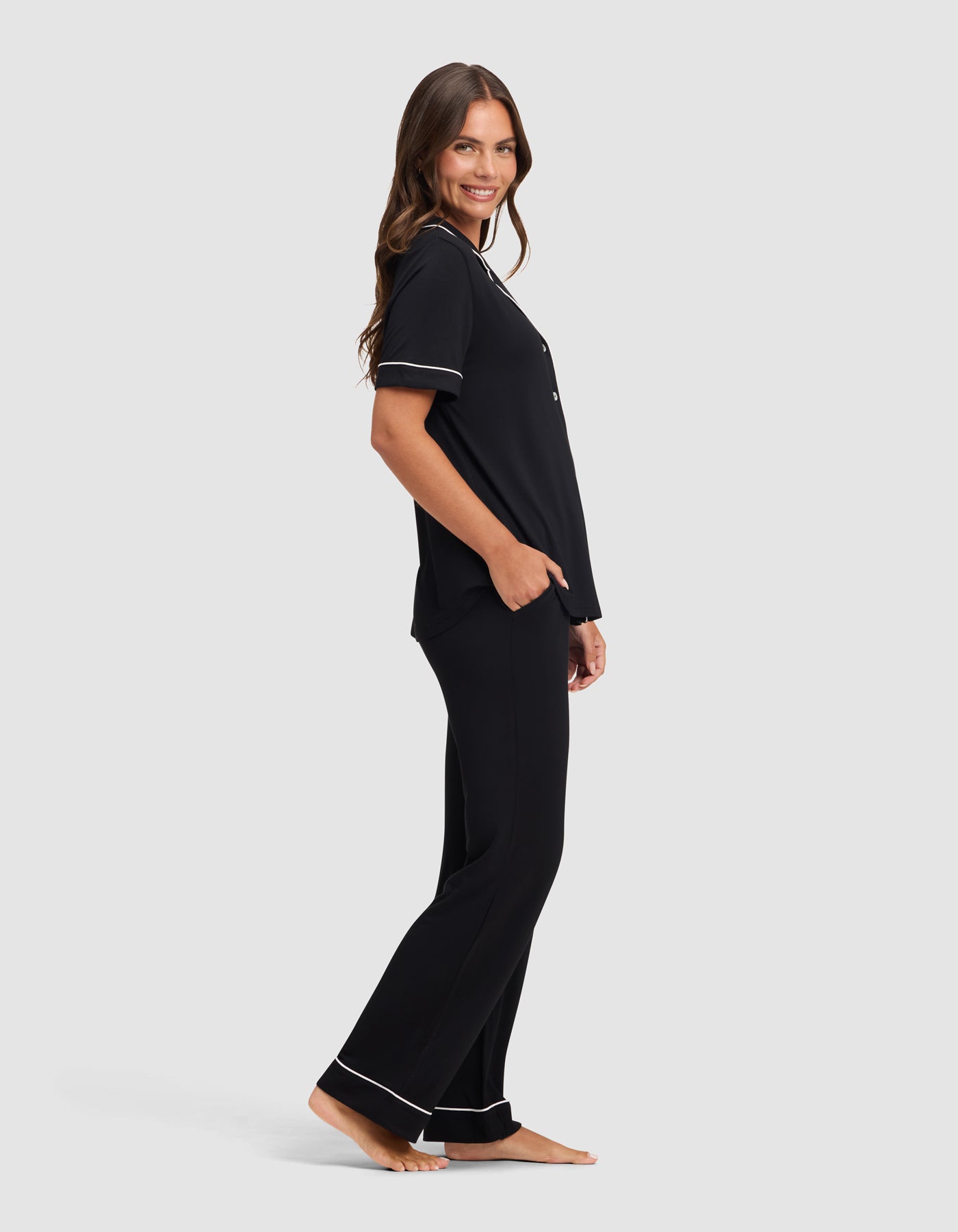 A woman with long brown hair poses barefoot and smiling against a light gray background, wearing Cozy Earth's Women's Bamboo Stretch-Knit Short Sleeve & Pant Pajama Set in black with white trim.|Color:Black