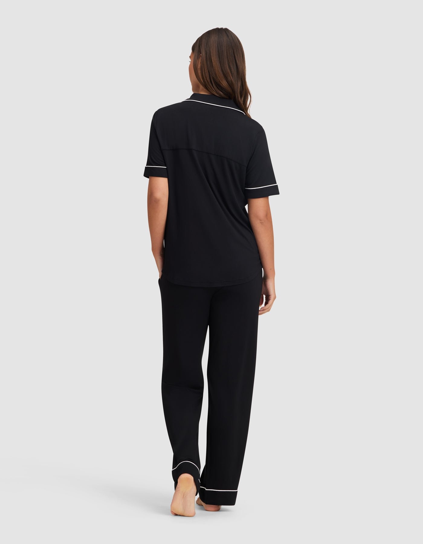 A woman stands barefoot facing away, wearing the Cozy Earth Women's Bamboo Stretch-Knit Short Sleeve & Pant Pajama Set in black with white piping, set against a plain light gray background.