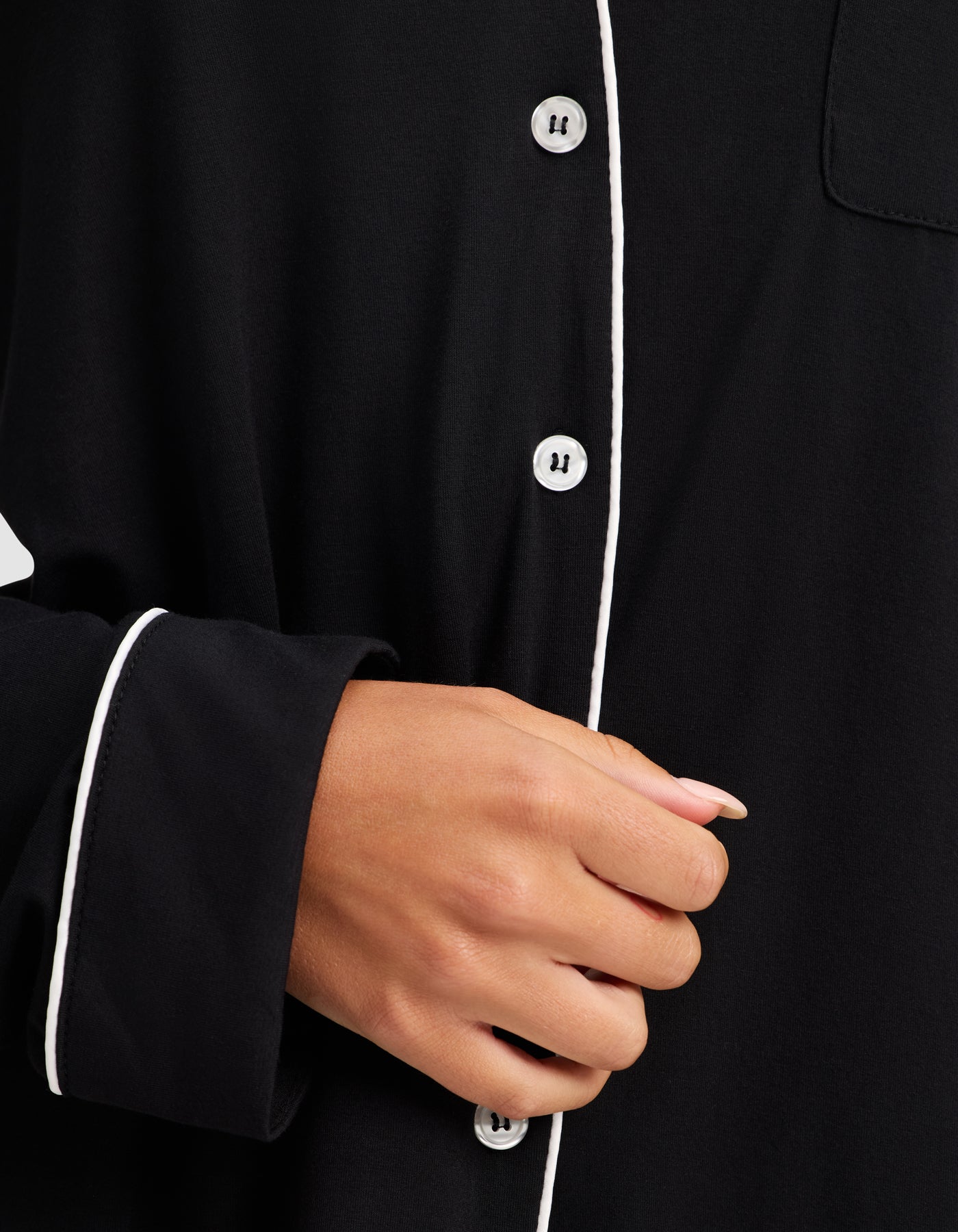 A close-up shows a hand holding the front of Cozy Earth's Women's Bamboo Stretch-Knit Long Sleeve Pajama Set, featuring white piping, white buttons, and a relaxed fit. The person’s nails are neatly manicured. 