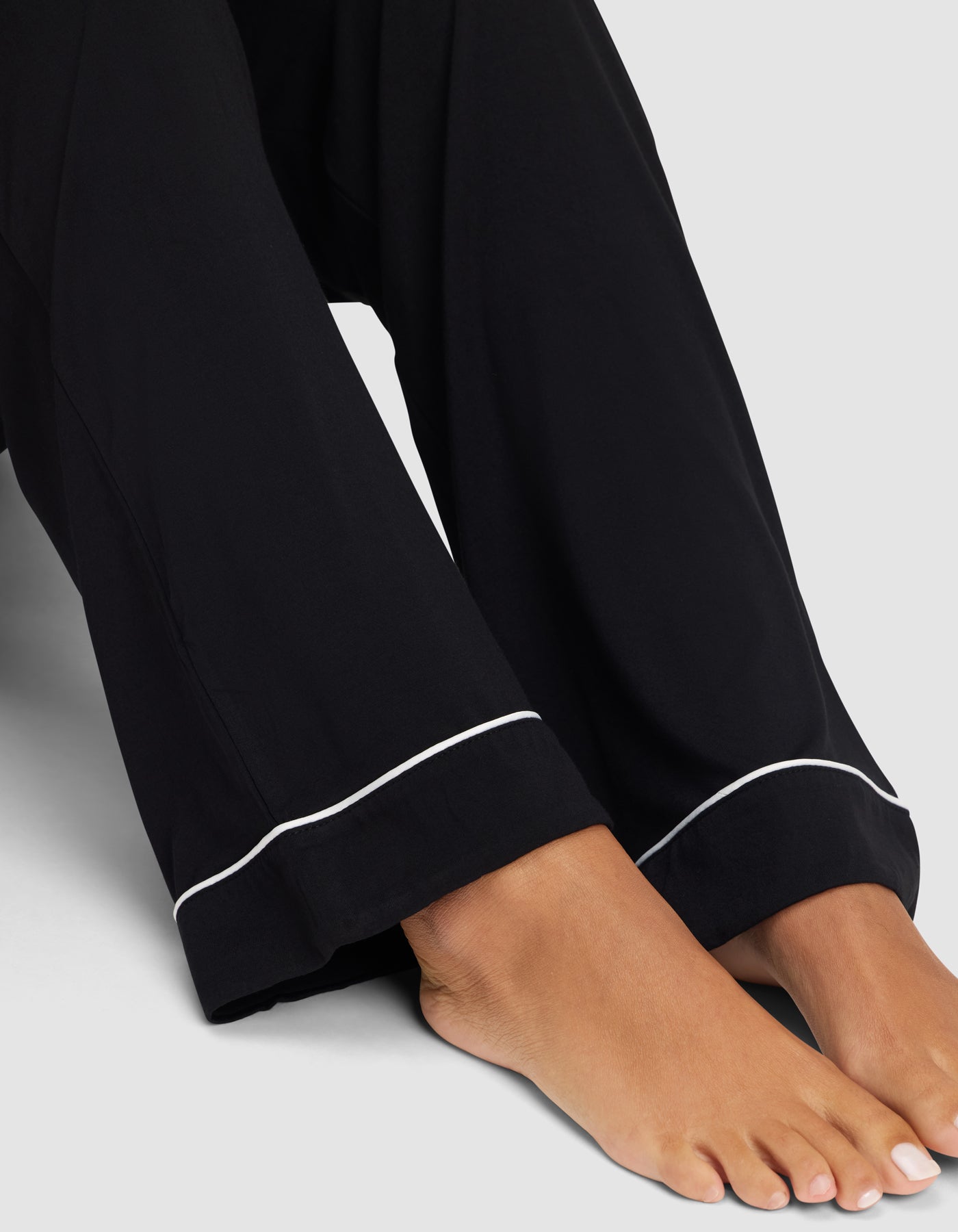 A close-up of bare feet and lower legs in Cozy Earth’s Women's Bamboo Stretch-Knit Long Sleeve Pajama Set, featuring loose black pants with white piping at the hem, set against a plain light gray background. 