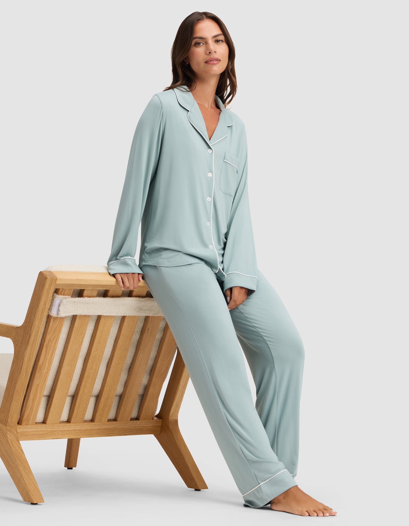 A woman in the Cozy Earth Women's Bamboo Stretch-Knit Long Sleeve Pajama Set in light blue sits casually on the arm of a wooden lounge chair with a neutral cushion, set against a plain light background.  