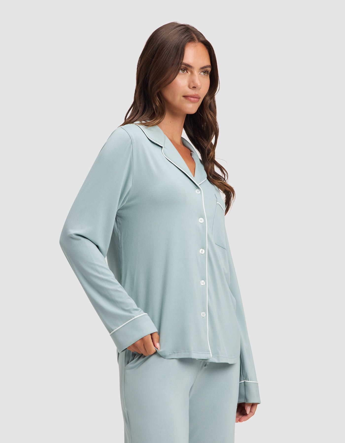A woman models the Cozy Earth Women's Bamboo Stretch-Knit Long Sleeve Pajama Set in light blue with white piping, one hand in her pocket, standing against a plain light background.  