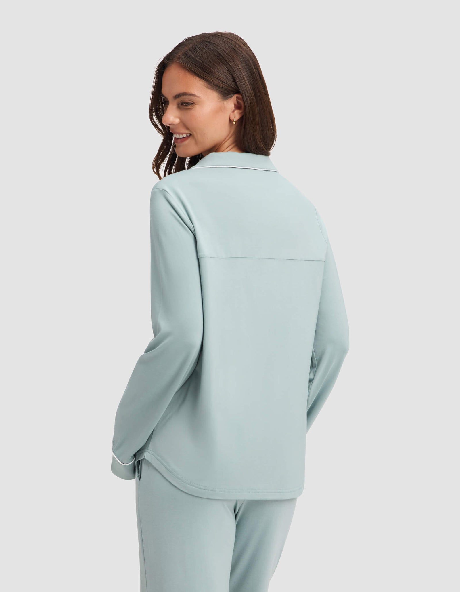 A woman with long brown hair wears the Cozy Earth Women's Bamboo Stretch-Knit Long Sleeve Pajama Set in light blue, standing against a plain white background and smiling over her shoulder.  