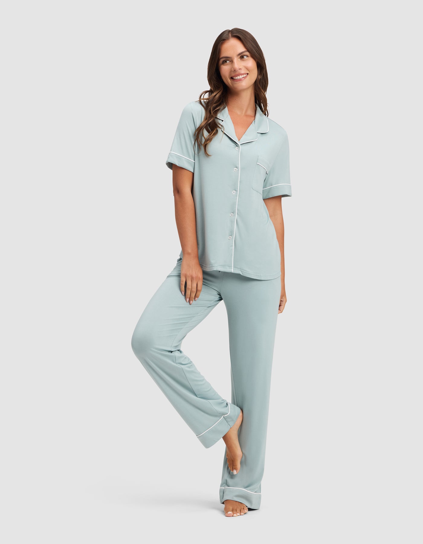 A woman with long brown hair smiles while wearing the Cozy Earth Women's Bamboo Stretch-Knit Short Sleeve & Pant Pajama Set, standing barefoot against a plain white background. 
